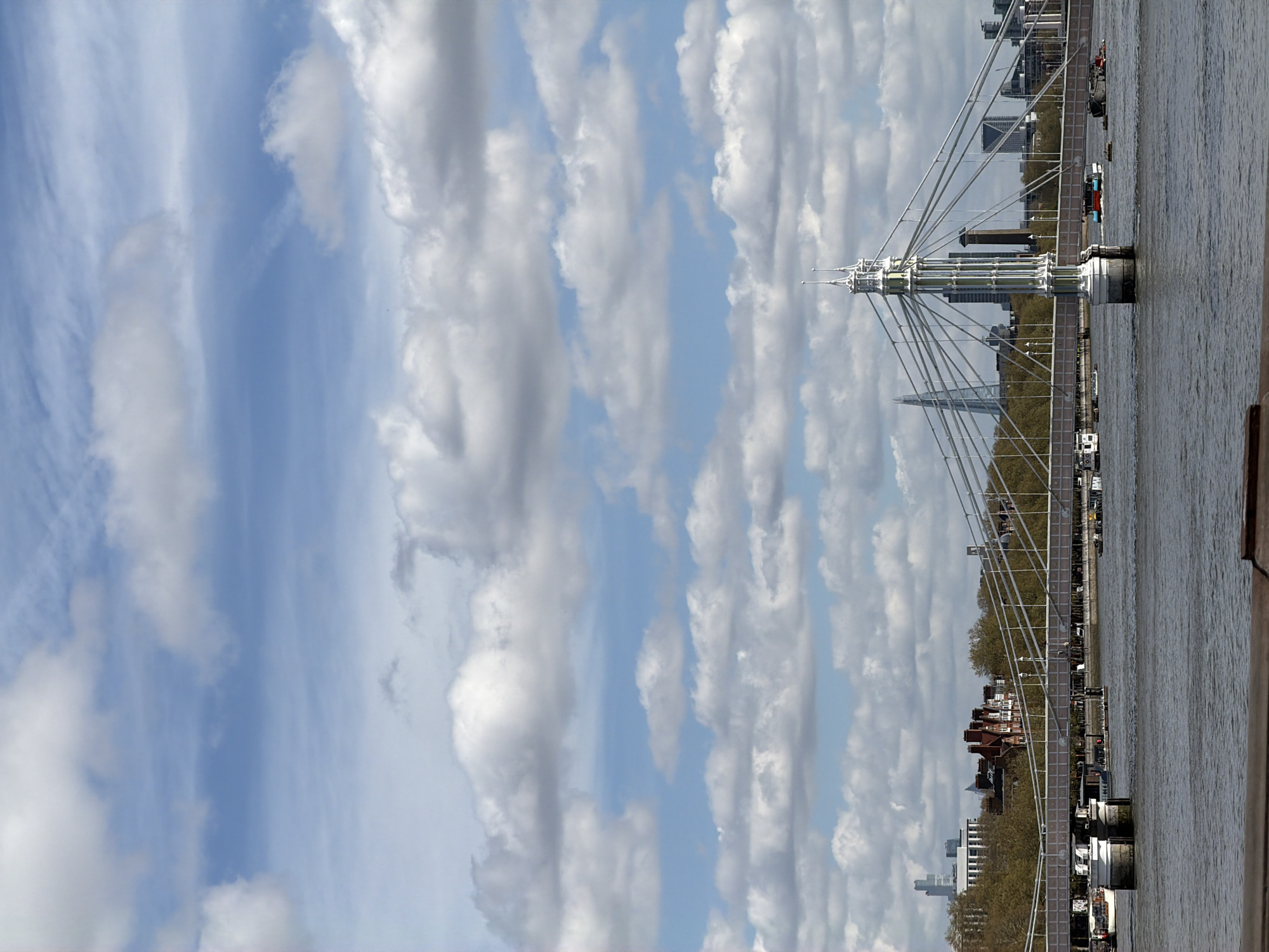 The image captures the Albert Bridge in London, UK, from a slightly low angle. The bridge is a prominent feature with its intricate design of suspension cables and white ornate towers. The River Thames flows horizontally across the right side of the image, with the bridge spanning over it. On the far bank, buildings are visible, including residential structures with red roofs and taller modern buildings in the distance. The sky above is filled with a mix of blue patches and puffy white clouds that cover most of the sky, adding a dynamic and airy feel to the scene. The image is taken during the day, as indicated by the ample natural light. The photograph is oriented sideways.