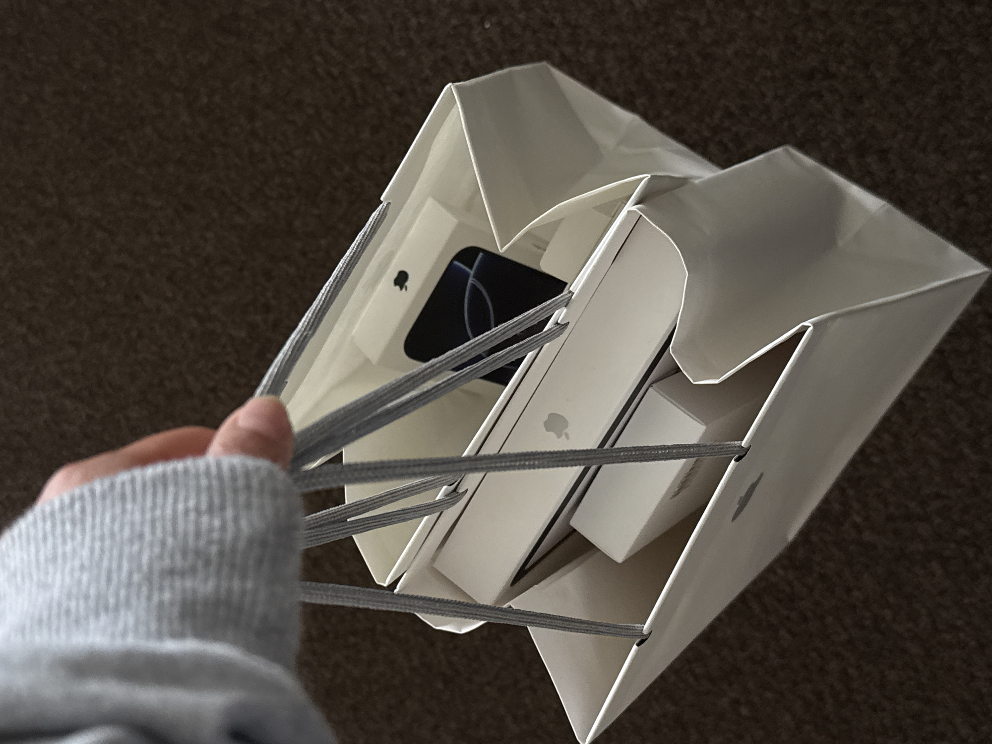 A person's hand, partially visible on the left, holds the gray string handles of two white shopping bags. The hand is covered by a gray, ribbed knitted sleeve. The perspective is looking down into the open tops of the bags.
Inside the shopping bags, several white boxes are visible, indicating recent purchases. In the bag closer to the viewer, a larger white box with the distinct Apple logo on its side is prominent. Adjacent to it is a smaller white box. The second bag, slightly behind and to the left, also contains white boxes. One of these boxes has its top flap open, revealing a dark-colored electronic device (possibly a smartphone or tablet) with a glowing screen displaying a white circular pattern. A smaller white box with an Apple logo is also visible deeper within this bag.
The background consists of a dark, textured surface, likely a carpet or rug, providing a muted contrast to the bright white bags and boxes. The lighting appears to be even, suggesting an indoor setting. The image captures the immediate aftermath of a shopping trip, with the new Apple products freshly placed in their bags. The only visible text/logo is the iconic Apple logo on several of the product boxes.