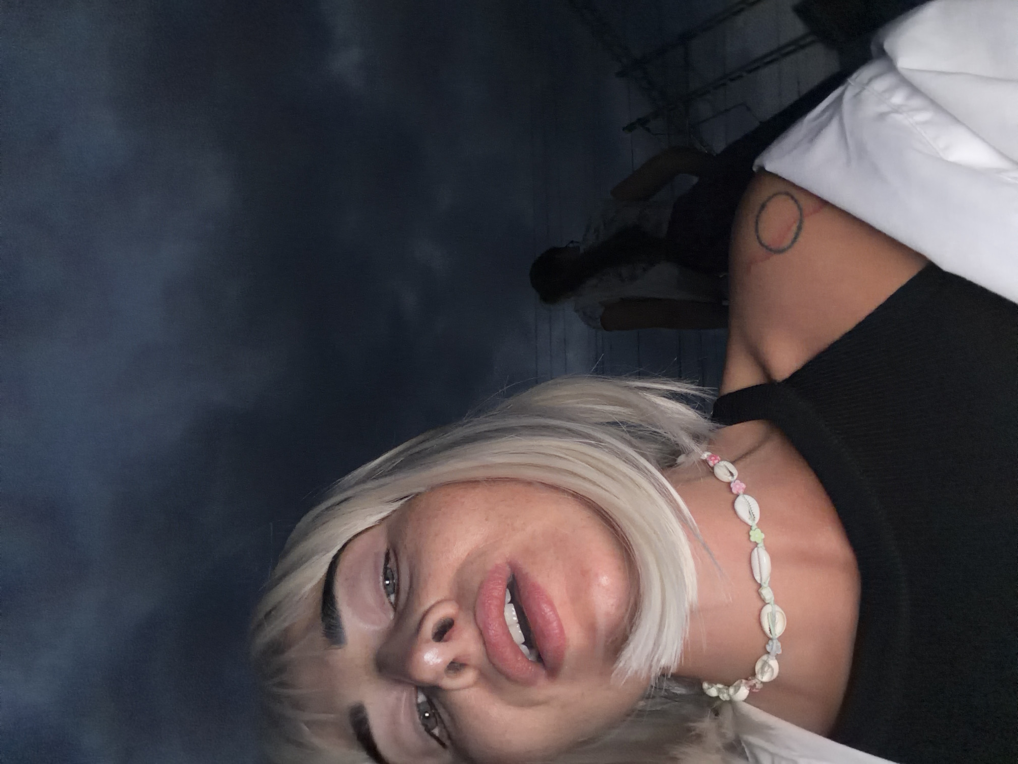 The image is a close-up selfie of a young woman with blonde hair and a seashell necklace. She is wearing a black tank top and a white shirt. Her lips are slightly parted, revealing her teeth. In the background, another person is visible, silhouetted against a dark, cloudy sky. The scene appears to be outdoors, possibly at dusk or night, given the low light conditions and dramatic sky. The presence of what appear to be power lines in the background suggests an urban or semi-urban environment. The photo was taken in Sochi, Russia.