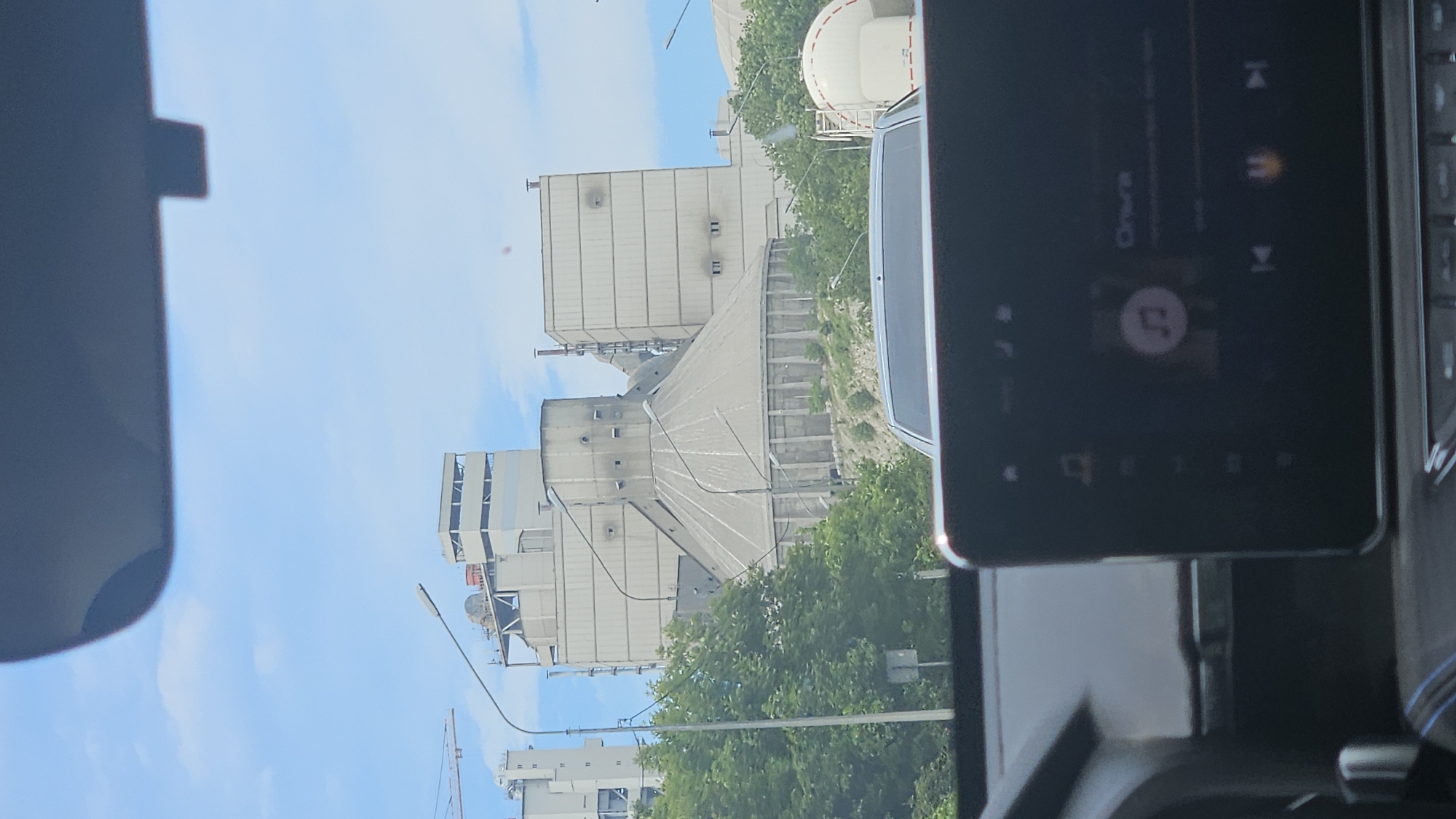The image, taken from inside a vehicle, shows a large industrial complex, likely a cement plant, situated in Verkhnebakanskiy, Russia. The plant features numerous concrete silos and processing structures, some with visible pipework and ventilation shafts. A large, conical structure with a ribbed exterior dominates the center of the complex. Trees line the foreground, obscuring the base of some of the industrial buildings. A white truck trailer with a red stripe is visible to the right of the complex. The sky above is blue with scattered clouds. In the foreground, the dashboard of the vehicle is visible, including a dark touchscreen display showing a music playback interface with album art and song title. The overall scene depicts an industrial landscape on a clear day, observed from a moving vehicle.