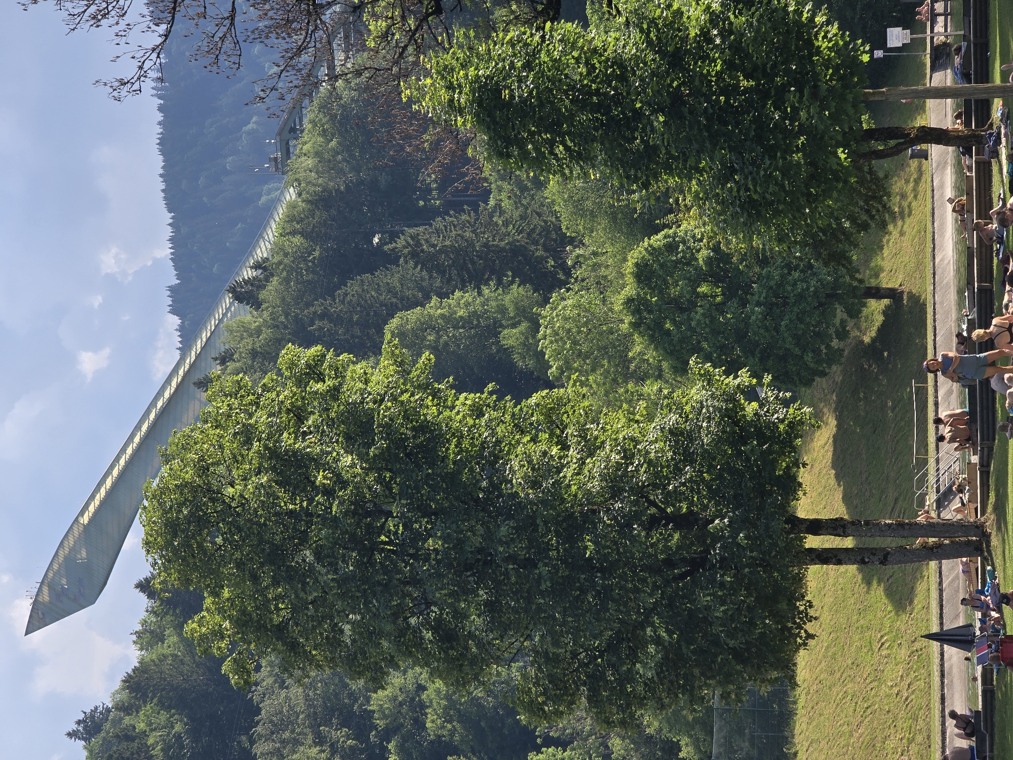 This is an outdoor scene in Garmisch-Partenkirchen, Germany, featuring a large ski jump structure in the background, a lush green forest, and a crowded swimming area in the foreground. The ski jump, a prominent architectural landmark, is angled upwards towards the sky, its sleek, modern design contrasting with the natural landscape. Several people are visible in the swimming area, some sunbathing on towels on the grass, while others are in or near the water, which appears to be a large pool or natural body of water. The trees are full of green leaves, indicating a summer or late spring day, and the sky is partly cloudy with sunshine casting shadows. The overall atmosphere suggests a recreational day in a scenic location.