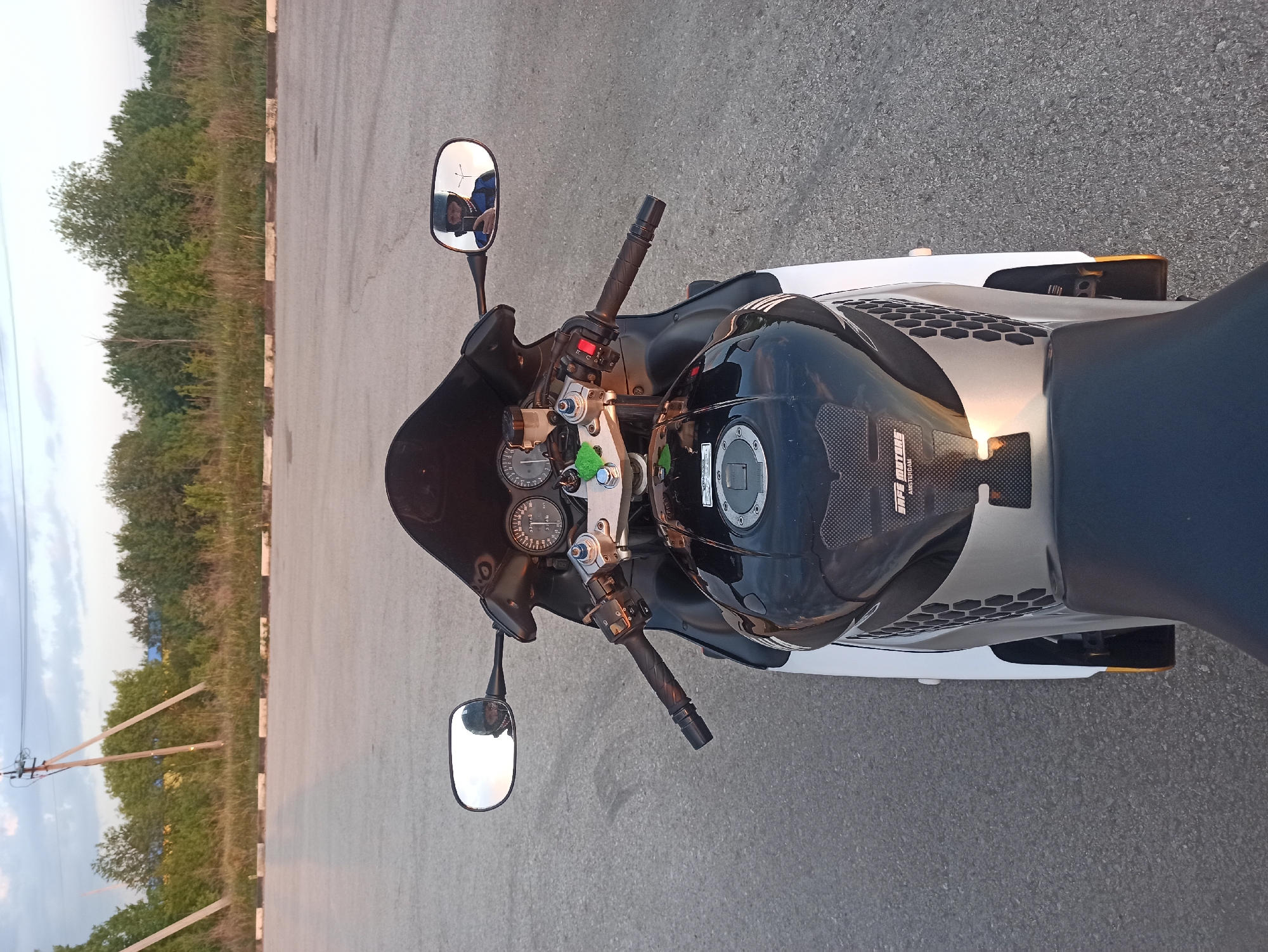 The image presents a close-up, top-down view of a motorcycle parked on an asphalt surface, possibly a road or parking area. The motorcycle has a black and silver color scheme with visible gauges and handlebars. A helmet rests on the fuel tank. In the motorcycle's right-side mirror, a reflection of a person holding a camera is visible. Beyond the asphalt surface, there is a curb followed by green trees. While it is not possible to know from this image alone if this is Perm, Russia, it is a city in Russia.