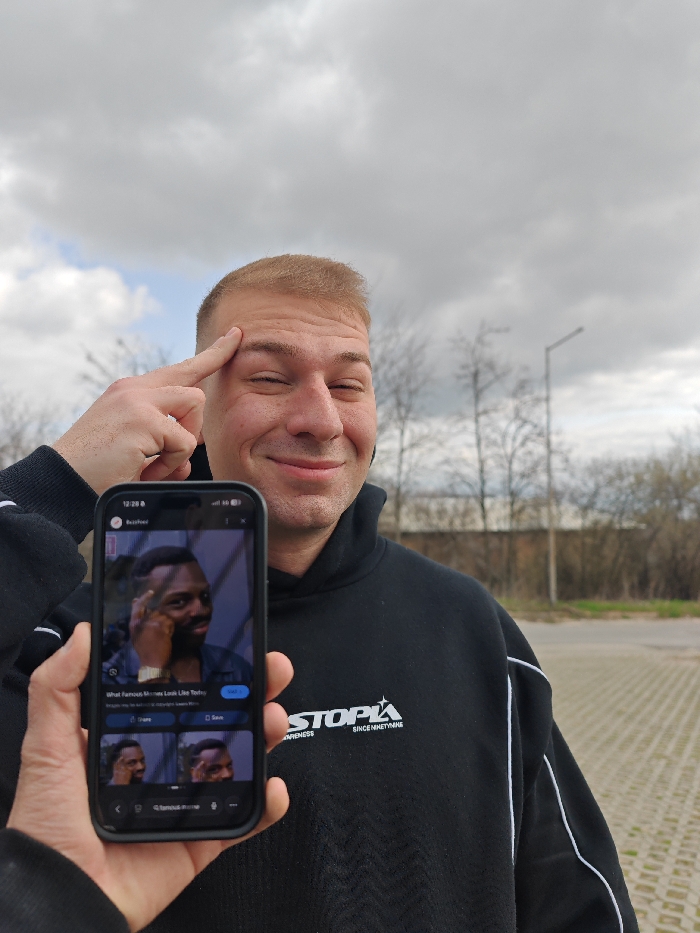 An outdoor medium shot captures a light-skinned male, estimated to be in his 20s or 30s, with short, light brown hair, posing in the city of Shumen, Bulgaria. He is smiling with closed eyes and has his left index finger pressed against his right temple. He wears a black hoodie featuring white piping along the sleeves and shoulders, and a white "STOPIA SINCE NINETYNINE" logo on the chest. In the foreground, a light-skinned left hand holds a black smartphone vertically. The phone's screen displays a clear image of the "Roll Safe" meme, depicting a man pointing to his head, along with what appears to be a Google image search interface with related thumbnails and the query "What Famous Memes Look Like Today." The background features a light gray paved surface, several bare trees, a dark, low-rise building or wall, and a gray street light pole, all under an overcast sky with diffuse clouds.