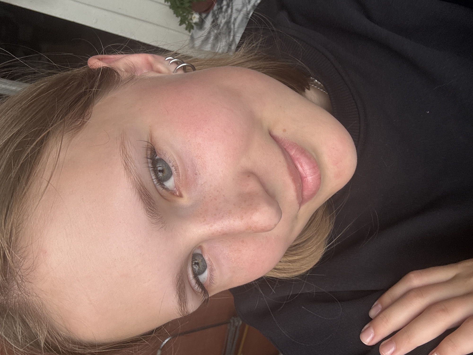 The image is a close-up, high-angle selfie of a young Caucasian female with fair skin, light brown hair, and green eyes. She has freckles scattered across her nose and cheeks. She is wearing a black sweatshirt and silver hoop earrings. Her left hand is visible, resting on her chest. The background appears to be outdoors with some foliage and a white structure. There is no indication that this image was taken in the city of Chaikovsky, Russia.