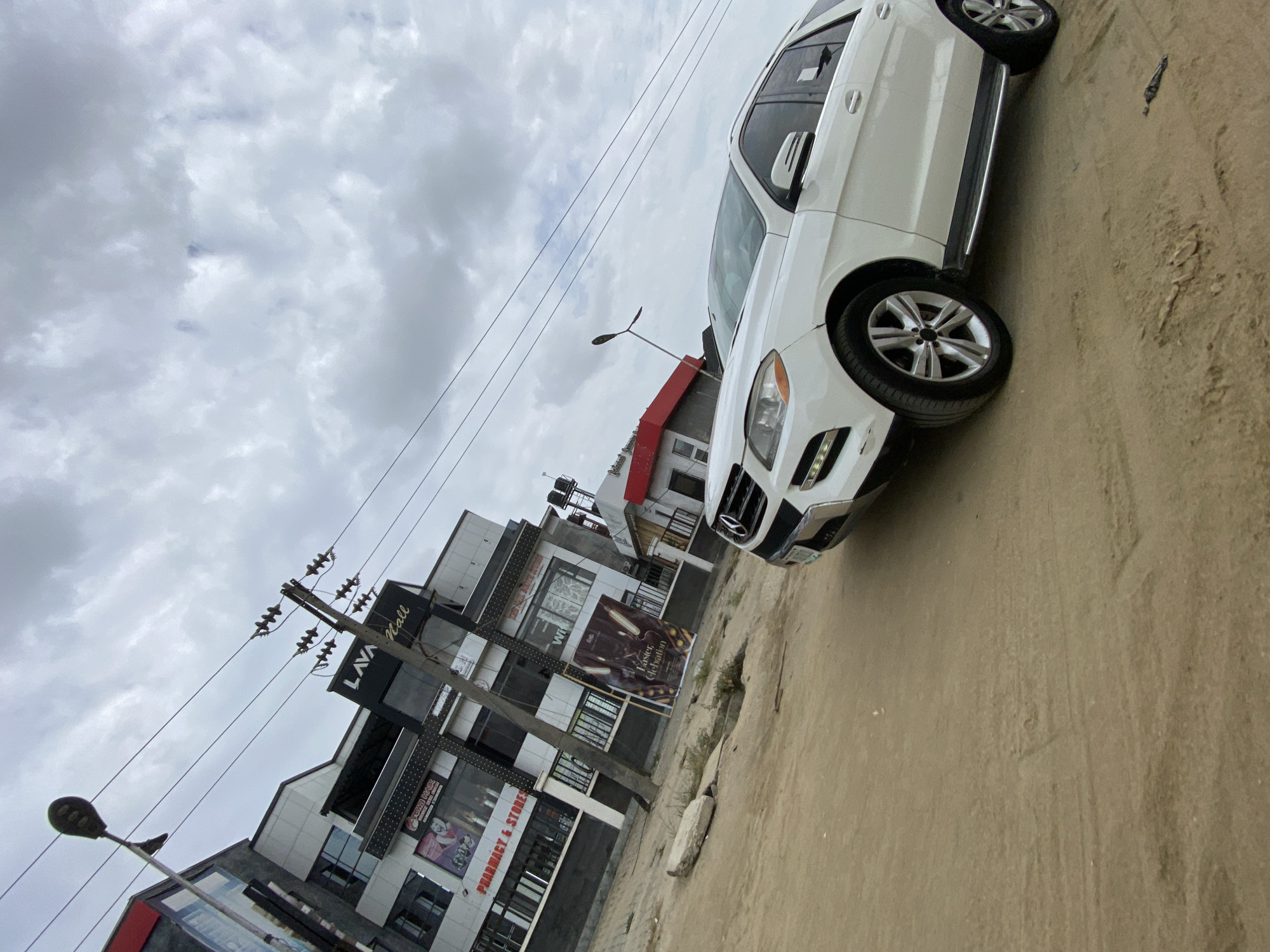 A slightly tilted eye-level shot captures a white Mercedes-Benz SUV parked on a dirt road in Ado, Nigeria. The car faces right. Behind the car, a multi-story commercial building with the sign "LAVA Mall" is visible. The sky is overcast. Power lines run diagonally across the upper left of the frame. A street light stands to the left of the building. The road surface is uneven and dusty.
