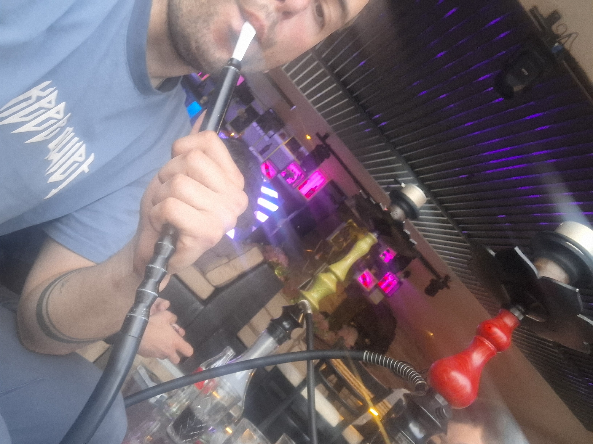 A person is smoking a hookah in what appears to be an entertainment venue with a low, dark ceiling featuring parallel lines of purple lights. The person is wearing a light blue t-shirt with white lettering. They are holding the hookah hose in their hand. Another person can be seen in the lower portion of the image. The hookah has a red base. The exact location within the city of Sofia, Bulgaria cannot be determined.