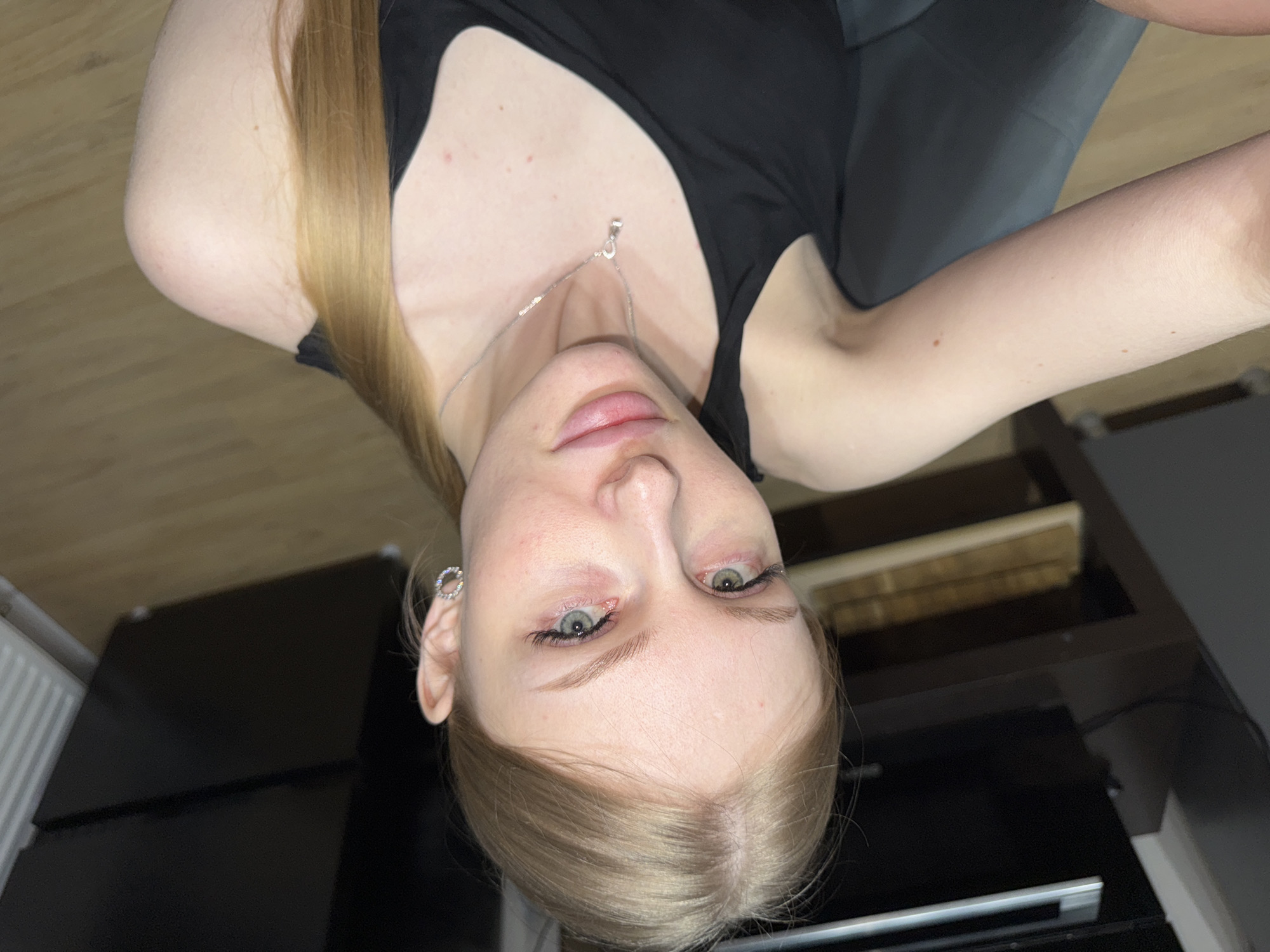 A fair-skinned woman with blonde hair is viewed from a close-up, tilted perspective. She is wearing a black tank top and a silver necklace. Her facial features are visible, including her light-colored eyes and pink lips. An earring is visible in her left ear. The background includes a dark gray surface, a light-colored wooden floor, and black cabinets or furniture. No location in the city of Perm, Russia is discernible.