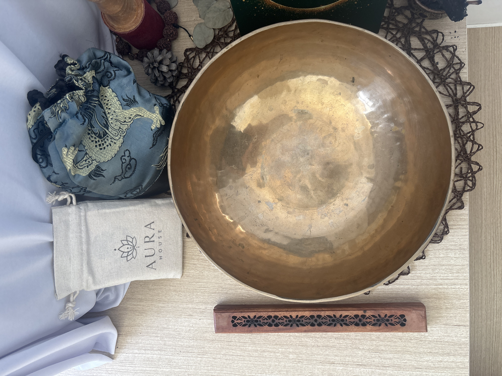 A still life arrangement is presented from a high-angle perspective, set against a light-colored wooden surface. In the foreground, a large, brass singing bowl dominates the center of the frame, its interior exhibiting a patina of age and use. To the left of the bowl, a rectangular wooden incense holder with intricate cutouts is positioned vertically. Above the bowl and slightly to the left, a small, light-colored linen pouch with a drawstring closure is visible. The pouch features a logo with a lotus flower and the text "AURA HOUSE." To the right of the linen pouch, a blue drawstring bag with a gold embroidered dragon motif is placed. Further to the right and behind the singing bowl, a dark red cylindrical object, possibly a spool, stands upright. A strand of brown beads, resembling rudraksha, encircles the base of the red object. Scattered around these items are a few pinecones and dried eucalyptus leaves. A white cloth is draped in the upper left portion of the image, and a dark green object with gold flecks is partially visible in the lower right corner. The lighting suggests an indoor setting, possibly with natural light contributing to the illumination. There are no people or animals present in the image. The overall composition suggests a curated collection of items related to meditation or spiritual practice.