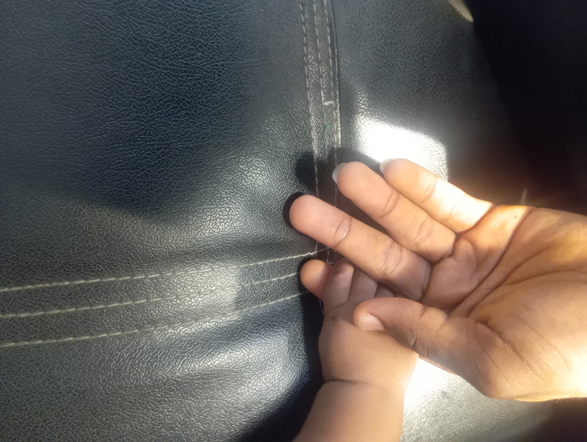 A close-up, high-contrast image depicts an adult's hand gently cradling or touching a baby's tiny hand or foot. Both individuals have dark brown skin tones. The adult's hand is visible on the right side of the frame, with distinct fingers and nails, while the baby's much smaller hand or foot, featuring undeveloped fingers/toes and nails, is positioned towards the bottom left.
The hands rest on a dark, textured surface, possibly leather or faux leather upholstery, which features prominent light-colored double-stitched lines running horizontally across the upper left and middle sections. Bright light, likely sunlight, streams in from the top right, creating strong highlights on the hands and the surface, particularly on the right side, and casting shadows that add depth to the composition. The lighting suggests it is daytime.
The scene conveys a sense of tenderness, connection, and protection between the adult and the infant. No text is visible in the image. The setting appears to be an interior space or a vehicle, given the nature of the upholstered surface.