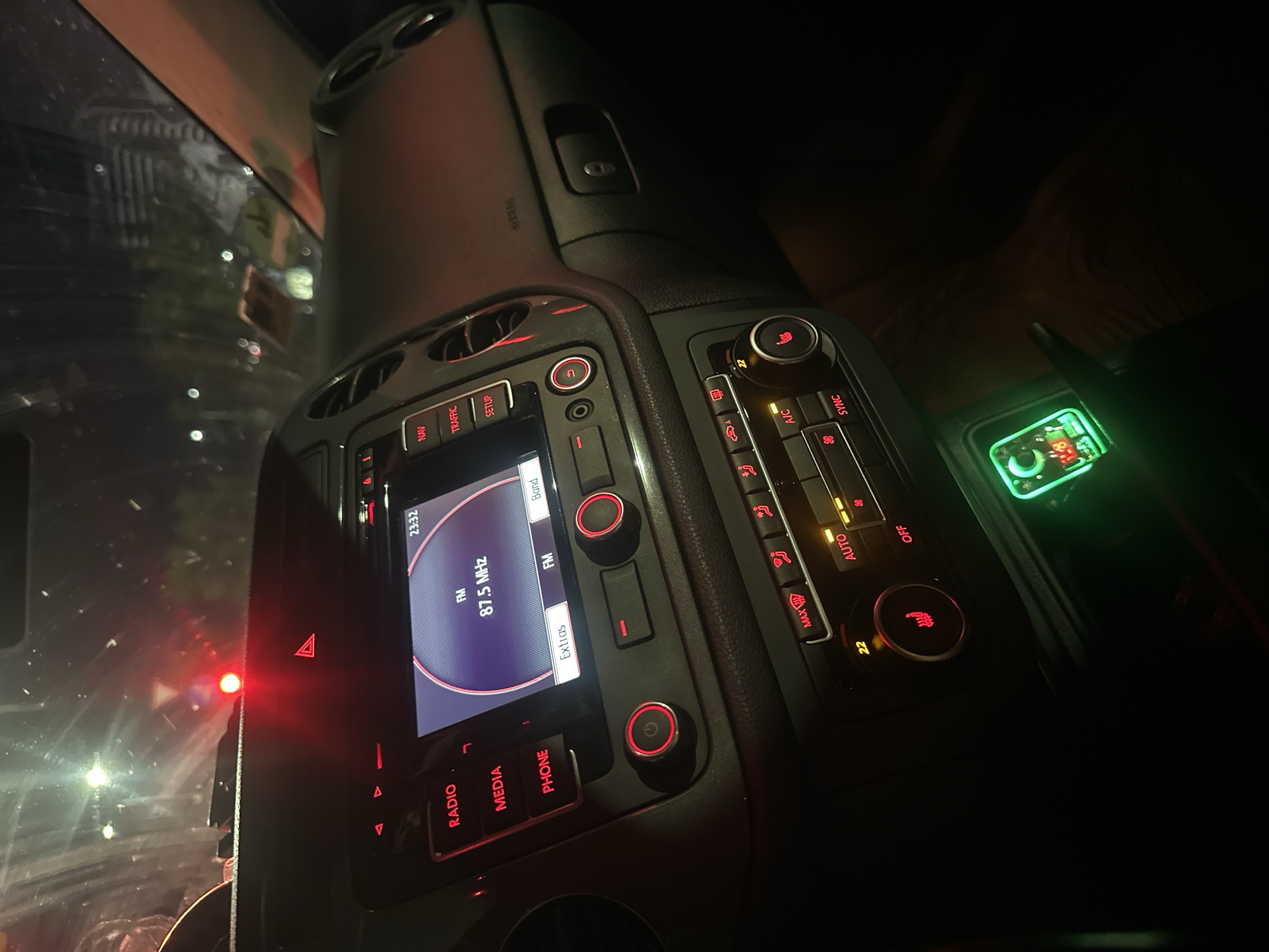 The image shows the interior of a car at night. The dashboard is illuminated by red and green lights from the car's controls and a small electronic device plugged into a socket. The radio display shows "FM 87.5 MHz". A red light is visible on the windshield. The car appears to be parked on a street in Sofia, Bulgaria, based on the overall context of the user's request. There are no people visible in the image.