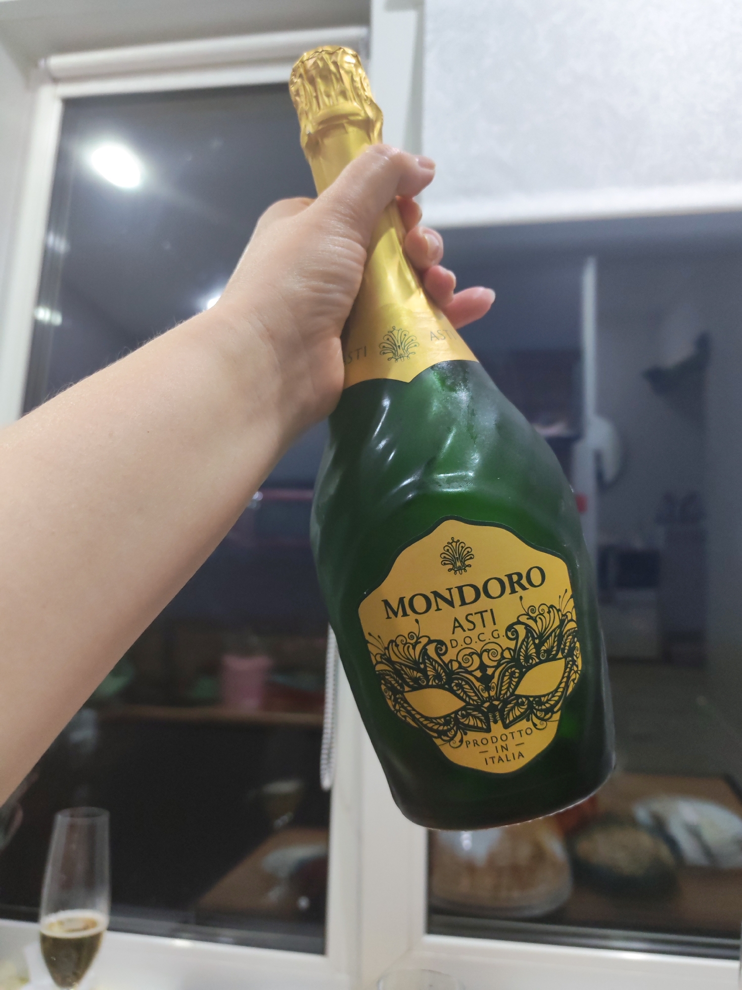 A fair-skinned person is holding a green glass bottle of Mondoro Asti DOCG sparkling wine. The bottle has a gold label with a stylized mask design and the words "PRODOTTO IN ITALIA." The bottle's neck is wrapped in gold foil. A partially filled champagne flute stands on a surface near the bottom left of the frame. The background appears to be an interior setting, possibly a kitchen, with a window behind the subjects, showing reflections of items and light. The location in the city of Izhevsk, Russia, is not discernible from this image.