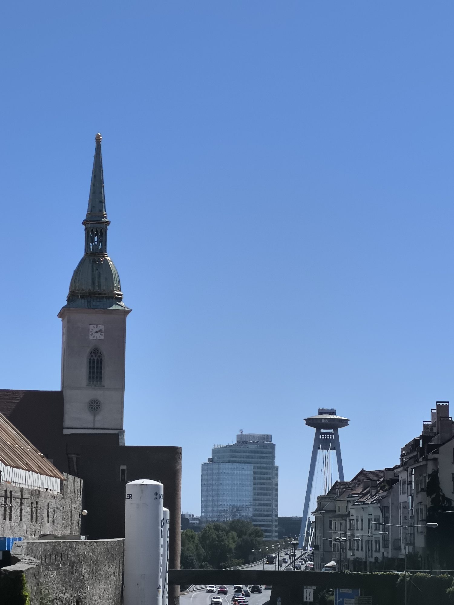 This is a photograph of the Bratislava cityscape under a clear blue sky. In the foreground, on the left, is the spire and upper portion of St. Martin's Cathedral, a prominent landmark in Bratislava. The cathedral has a tall, slender green spire with a golden pinnacle, and a grey stone tower with a clock face and gothic-style windows. To the right of the cathedral, a modern glass skyscraper is visible. Further in the background, the UFO Observation Deck, an iconic structure of the Most SNP (Bridge of the Slovak National Uprising) bridge, stands tall. The bridge, a cable-stayed bridge, spans across a body of water, with traffic visible on it. To the far right, a row of traditional European buildings with pitched roofs lines a street. The scene captures a mix of historical and modern architecture, typical of Bratislava, the capital of Slovakia. There are no people clearly visible in the photograph. The environment appears to be an urban setting on a sunny day.