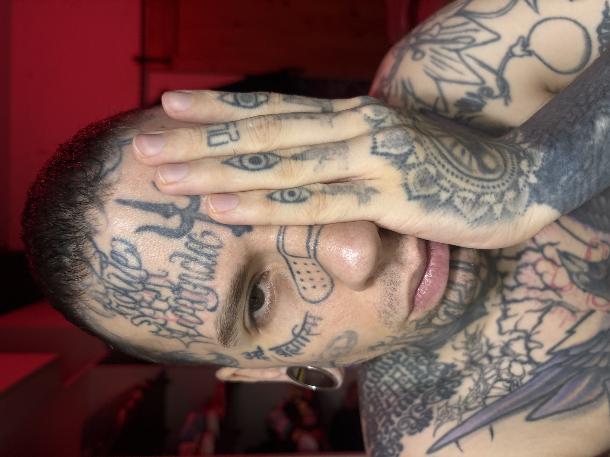 A man with extensive tattoos on his face, head, neck, and arms is covering one eye with his hand. The tattoos include lettering, eyes, and other designs. He has an ear stretching. The background is partially obscured and appears to be indoors with red lighting. There is no clear indication of the location being in Buenos Aires, Argentina.