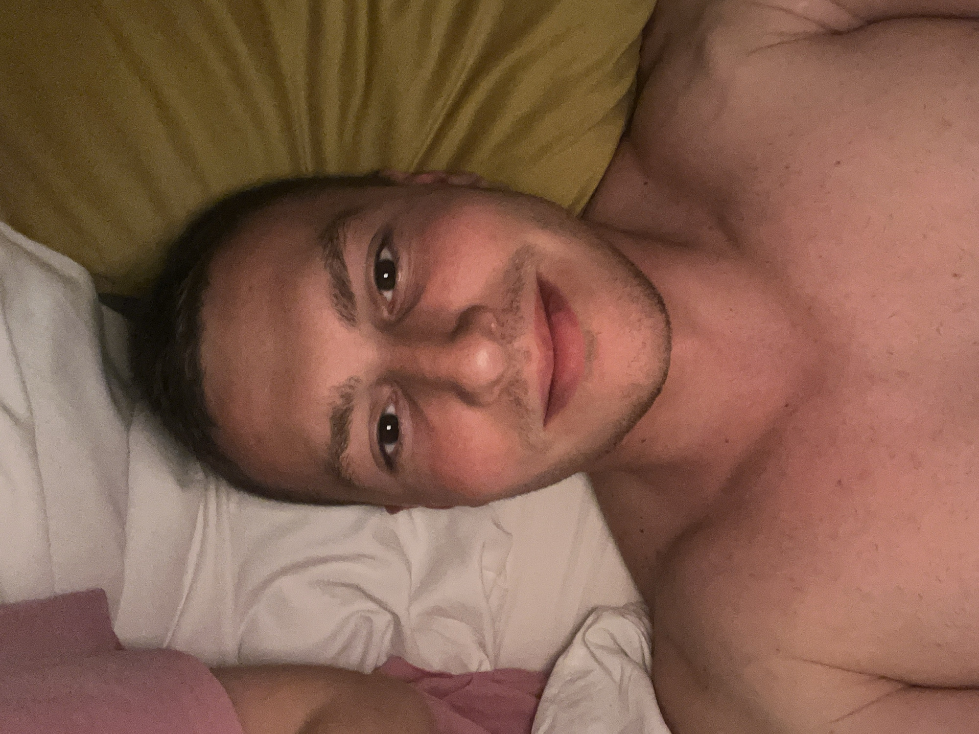 A man is depicted lying horizontally on a bed, looking directly at the camera. He has short dark hair and light stubble on his face. He is shirtless, revealing his upper chest and shoulders. A gentle, relaxed smile is visible on his face, and his eyes are open. His head rests on a white pillow, with a crumpled yellow pillow visible behind it. White and a pinkish-red colored bedding are also seen. The lighting in the room is dim and warm, suggesting an indoor setting, likely in the evening or at night. There is no visible text in the image.
