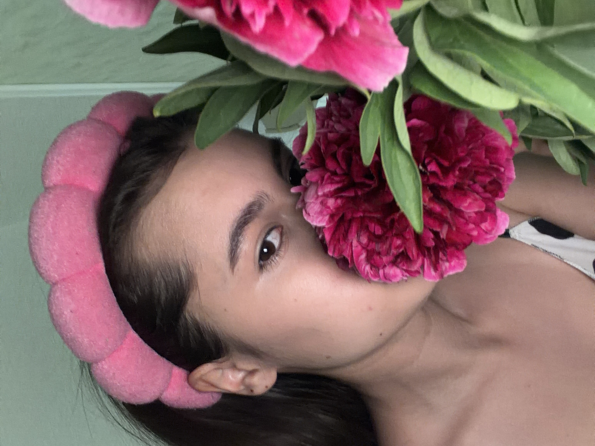 A young woman is positioned close to the camera, with a large, vibrant pink flower held near her face, partially obscuring her nose and cheek. She wears a pink, puffy headband. Her dark hair is visible around the headband and her shoulder. Her left eye is visible and appears to be brown. A portion of what appears to be a white garment with black spots is visible on her shoulder. The background is a light, neutral color. The exact location in Chaikovsky, Russia cannot be determined from the visual information provided.