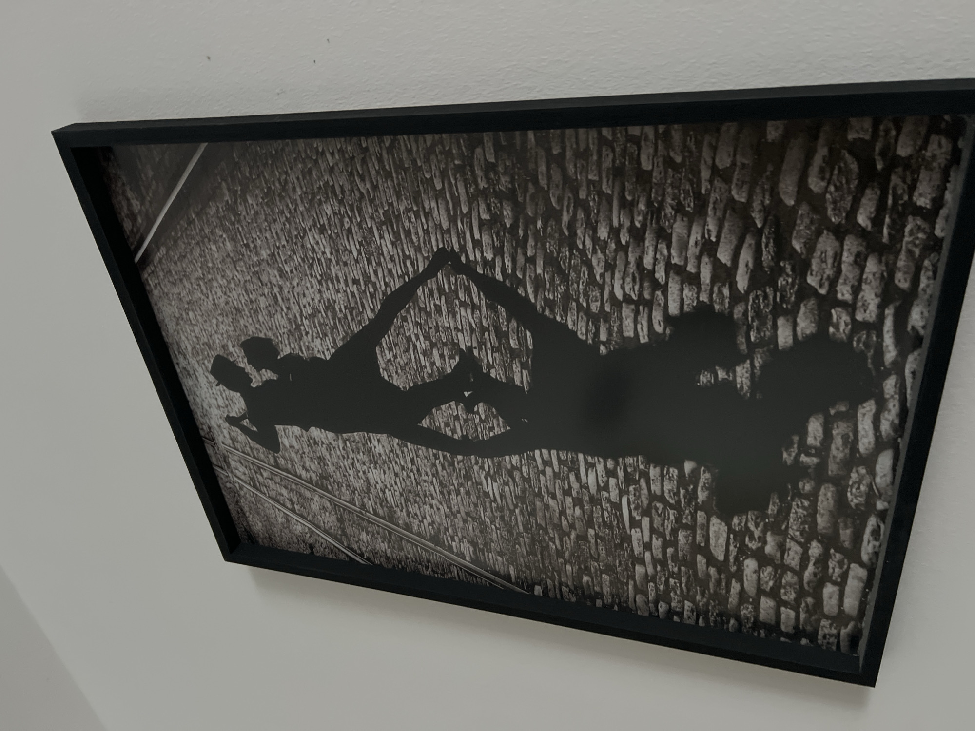The media file displays a framed black-and-white photograph hanging on a plain white wall, viewed from an angled perspective. The photograph's central subject is the elongated shadows of two individuals cast upon a textured cobblestone surface, likely a street or alleyway. The figures' shadows are silhouetted against the bright cobblestones, with their upper bodies and arms arranged to form a distinct heart shape, suggesting a moment of affection, romance, or connection. The extreme length of the shadows indicates a low sun angle, implying either early morning or late afternoon/evening. Faintly visible running along the cobblestones are two parallel lines that resemble train or tram tracks. The general setting, with its cobblestone streets and potential tracks, is characteristic of parts of Buenos Aires, Argentina. No discernible text is visible within the image.