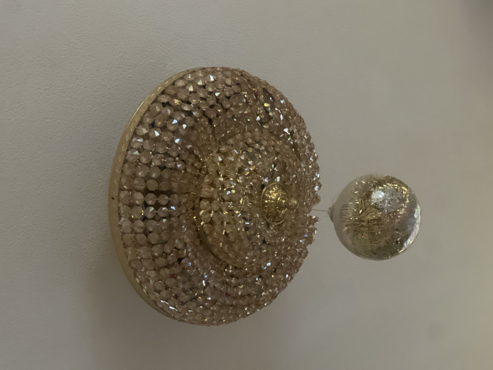 A close-up, overhead shot captures a decorative crystal chandelier against a neutral ceiling. The chandelier has a gold-toned base adorned with multiple layers of faceted amber-colored crystals, reflecting light and creating a shimmering effect. Hanging from the center of the chandelier is a clear glass orb filled with golden tinsel. The orb is suspended by a thin wire. The scene is illuminated by the ambient light of the room, casting soft shadows. The location is the city of Unknown, Unknown.