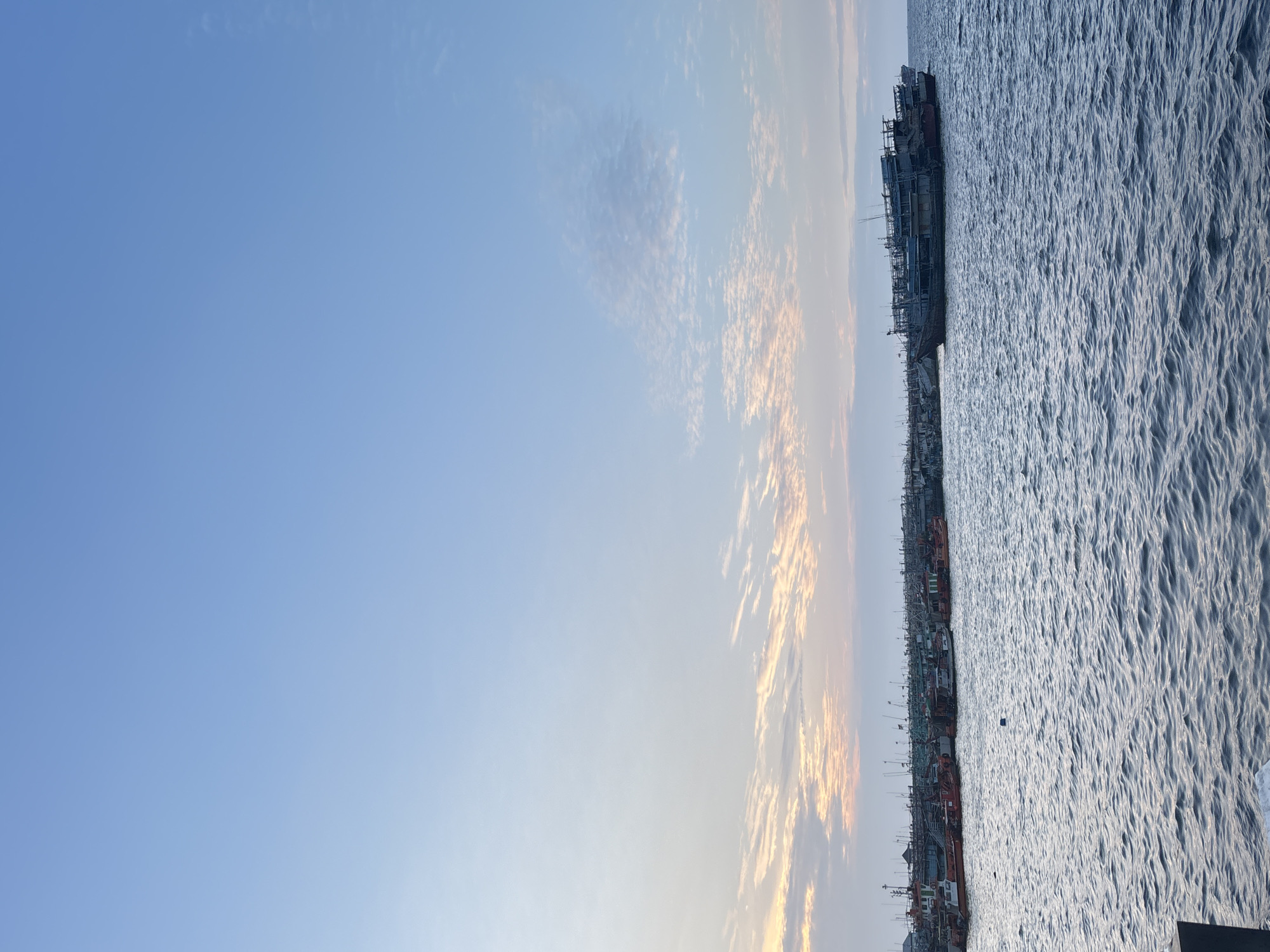 The image depicts a harbor in North Jakarta, Indonesia, at sunset. The sky is a gradient of soft blues and oranges, with wispy clouds scattered across the horizon. The calm sea reflects the muted colors of the sky. A fleet of fishing boats, varying in size and color, are moored together in the foreground and midground. The boats are densely packed, with many appearing to be older vessels with intricate rigging and multiple decks. Some boats are painted red and white, while others are darker hues. A larger vessel, appearing more modern and with a prominent superstructure, is positioned slightly to the right of center in the midground. The overall impression is of a working harbor at the end of the day, with the boats resting after a day's work.