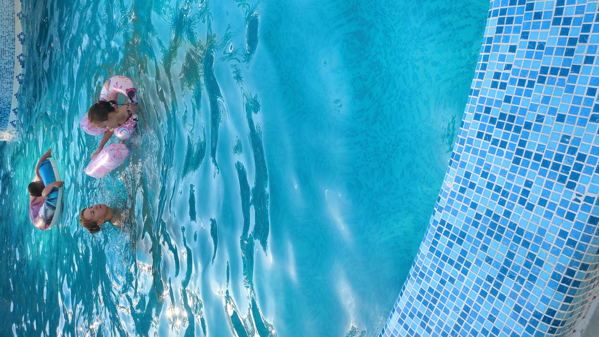 In a bright outdoor swimming pool, three individuals are visible. In the foreground, a woman with blonde hair pulled back is in the water, looking to her left. To her right, a young girl wearing a swimsuit and a pink inflatable ring around her torso is reaching out with her left hand. In the background, a child lies on a colorful inflatable ring in the water. The pool has a blue tiled edge and the water is clear and blue, with sunlight reflecting on its surface. The scene is consistent with a recreational area in Vadul lui Voda, Moldova.