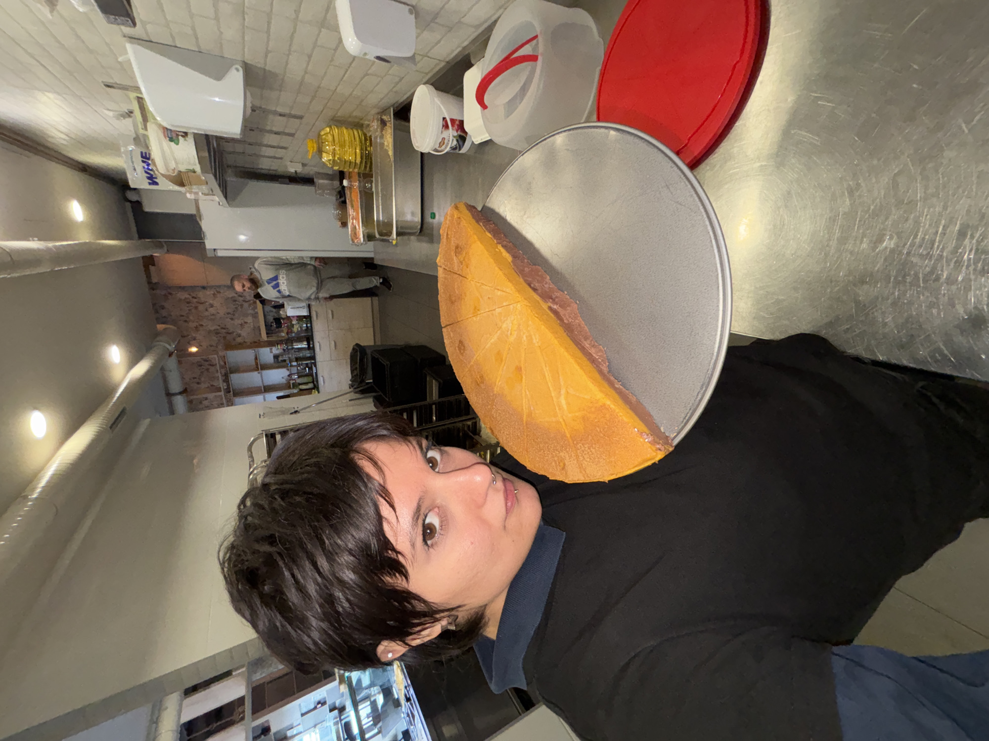 A person with dark hair and brown eyes wearing a black shirt and a blue undershirt is holding a round metal tray with a large piece of orange cheese cut into wedges on it. The photo appears to be taken in a commercial kitchen with stainless steel countertops and shelving visible in the background. There are various containers and kitchen equipment nearby. A second person is visible further in the background, standing near a refrigerator. The location appears to be in Sofia, Bulgaria.