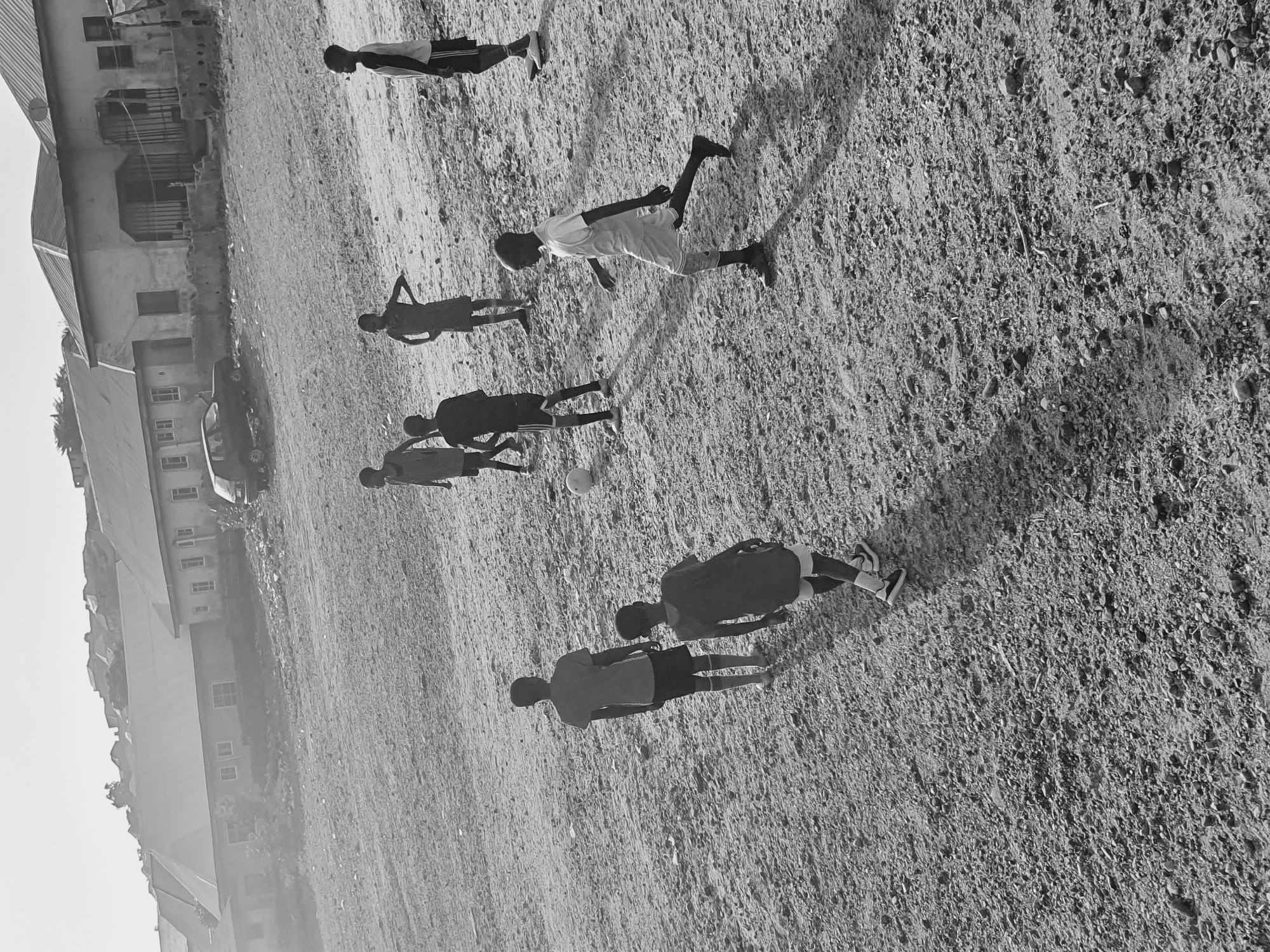 A black and white image shows six people playing soccer in a field near a building. The individuals are scattered across a dirt or grass field. Their clothing appears to be shorts and shirts, and their limbs are extended as if in motion. A soccer ball is visible near the center of the image. The building, including roofing and windows, is on the left of the image. This is likely Lokoja, Nigeria.