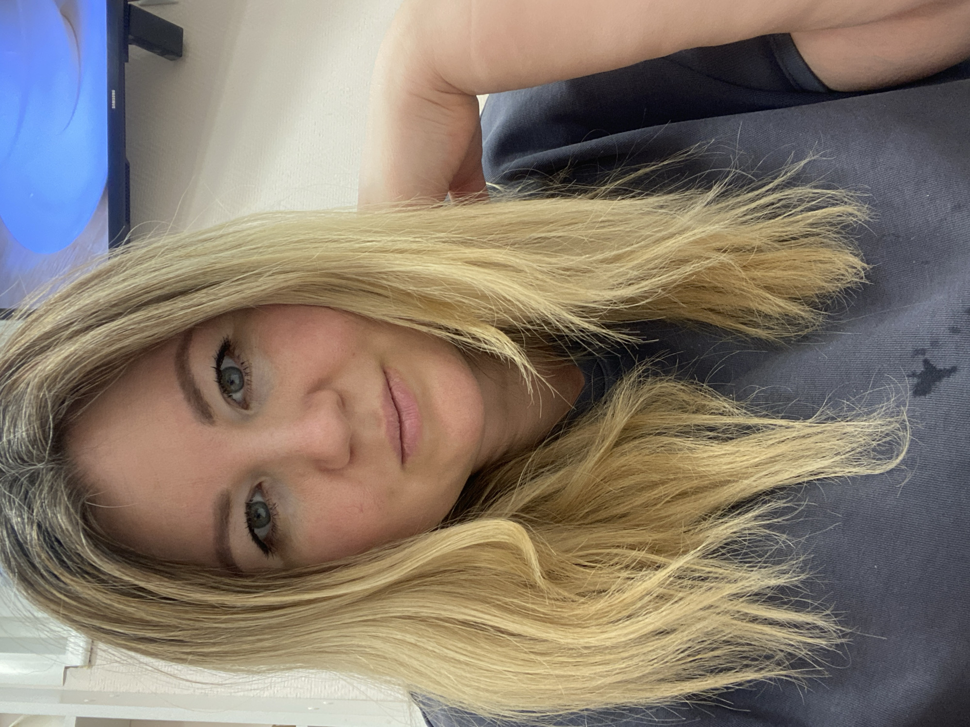 This close-up portrait, taken in Chaikovsky, Russia, features an adult woman with fair skin, light blonde hair, and blue-green eyes. She looks directly at the camera with a neutral to slightly pleasant expression, her lips closed in a subtle smile. Her long, blonde hair, which appears to be highlighted or dyed with some darker roots visible, cascades around her shoulders and face. She is wearing a dark grey or blue t-shirt or sweatshirt.
The setting appears to be indoors, likely a home or office. In the background, to the upper left, a portion of a wall-mounted flat-screen television is visible. The TV screen displays a bright blue, abstract image or background. The "SAMSUNG" logo can be clearly seen on the TV's frame. The surrounding walls are plain white or light-colored. The image is taken from a slightly upward angle, with the woman's head tilted. The overall impression is a casual, personal photograph.