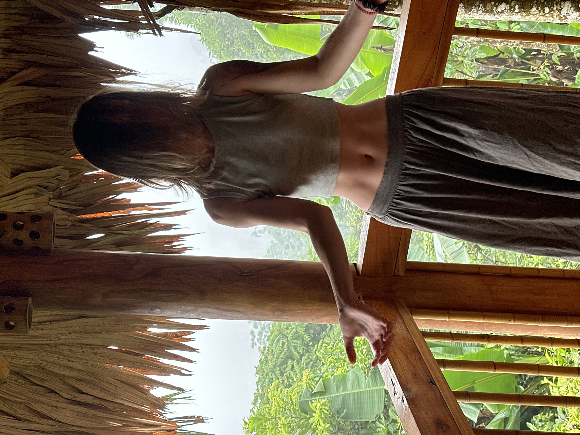 A person stands on a wooden balcony in Pù Luông Commune, Vietnam, their face obscured by their hair. They are wearing a grey cropped tank top and a long, grey skirt. Their arms are extended outwards, with their palms facing up. The balcony is covered with a thatched roof made of dried palm leaves. In the background, lush green trees and foliage are visible, with a misty atmosphere suggesting a humid or rainy day. The balcony railing is made of wood and bamboo poles.