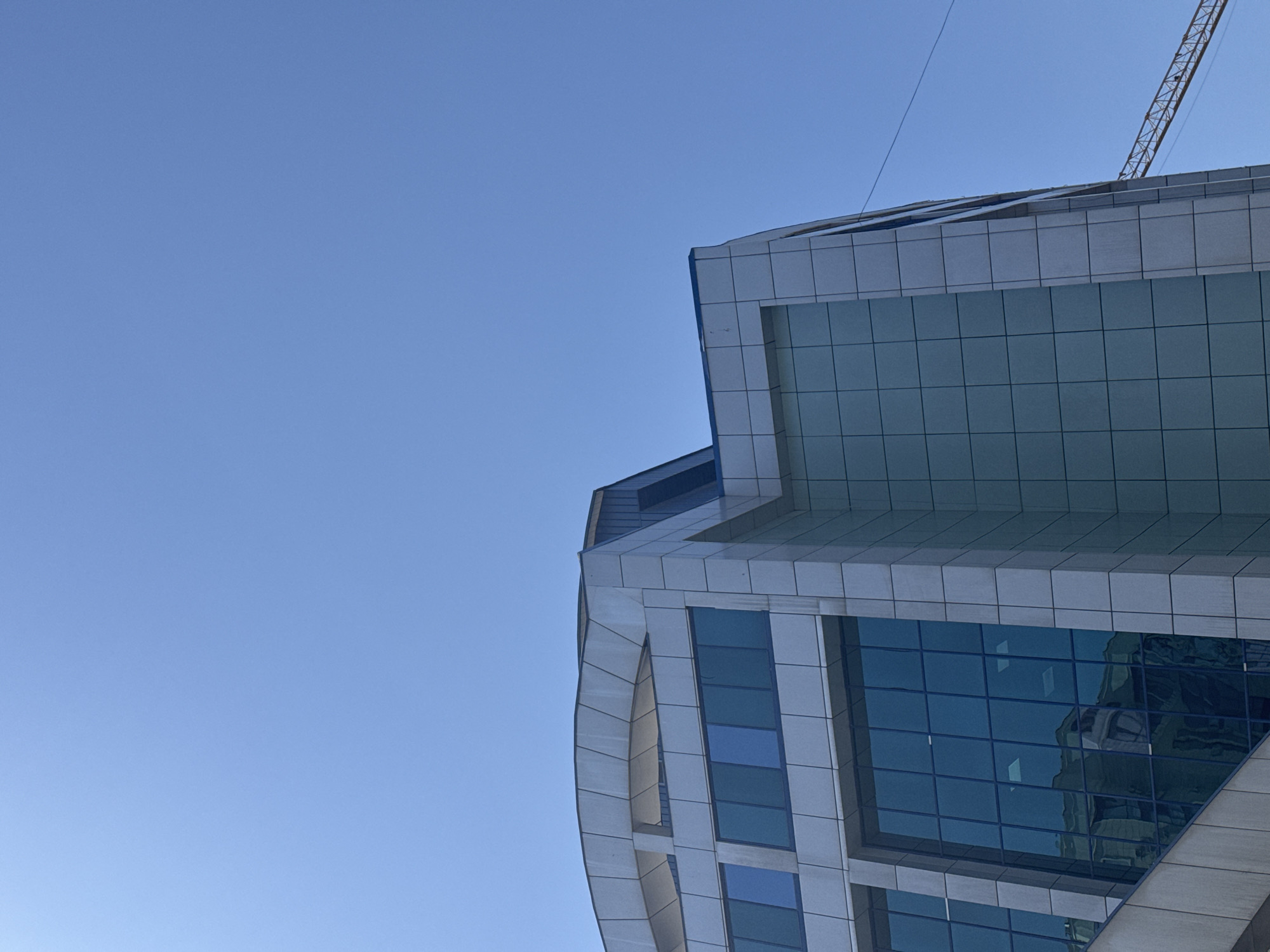 The image shows the exterior of a modern building with a glass facade and light-colored cladding. The building has several floors with large rectangular windows. The sky is clear and blue. In the background, a construction crane is partially visible. The scene appears to be in an urban environment.