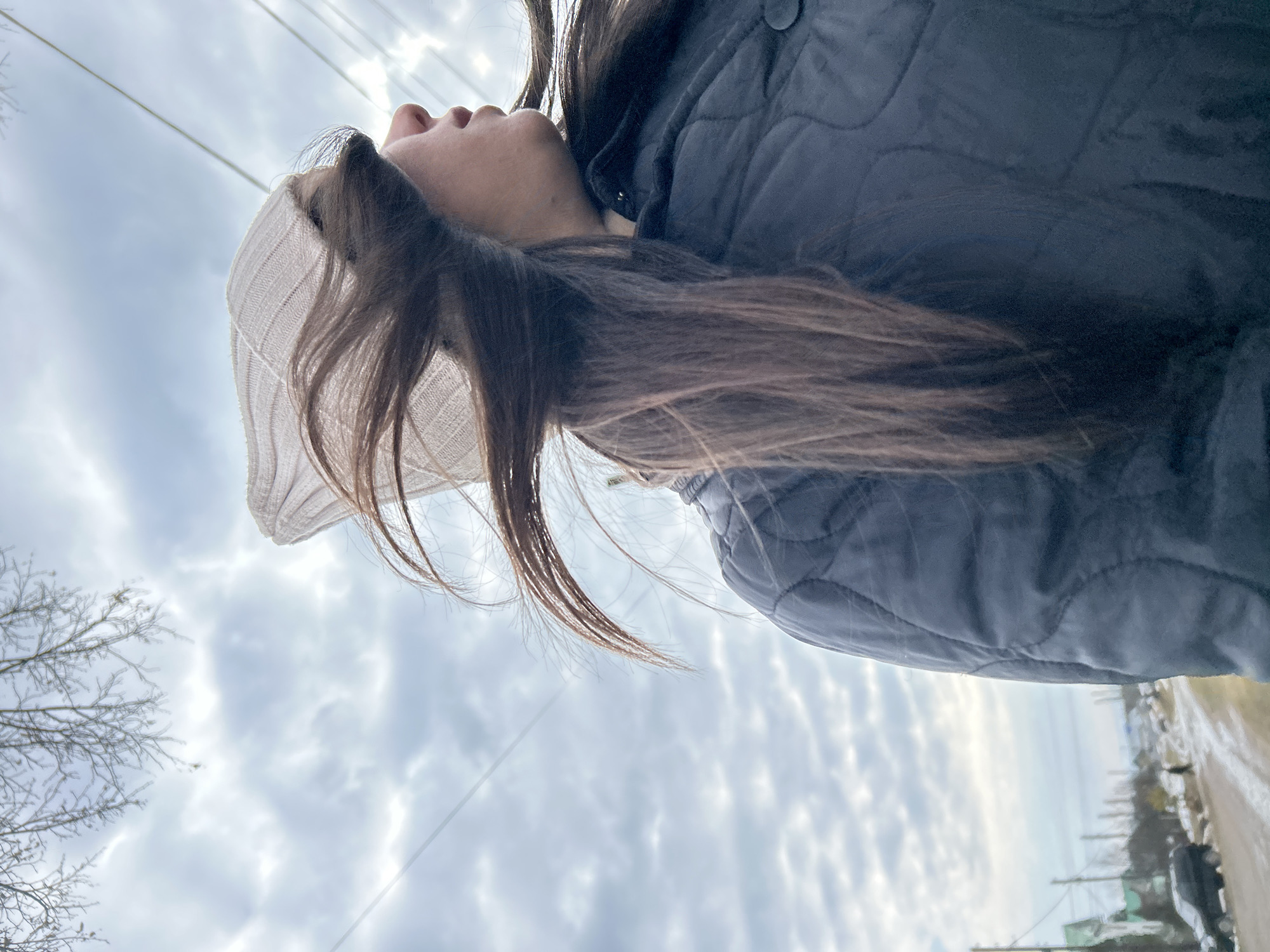 A low-angle photograph shows a person looking upward, with their face tilted towards the sky. They are wearing a light-colored knit hat and a dark, quilted jacket. The person has long, brown hair that is partially obscuring their face.
The background consists of a cloudy sky and elements of a street scene. Bare tree branches are visible in the lower-left corner of the frame, and power lines stretch across the sky. The street appears wet, and buildings are visible in the distance, suggesting an urban environment.
Based on the presence of Russian infrastructure (power lines, building styles), and referencing information available publicly, the location is likely in Chaikovsky, Russia, as requested. The scene appears to be shot during the day, but overcast weather conditions prevail. The person's expression is not visible, but their posture suggests contemplation or observation of the sky.
