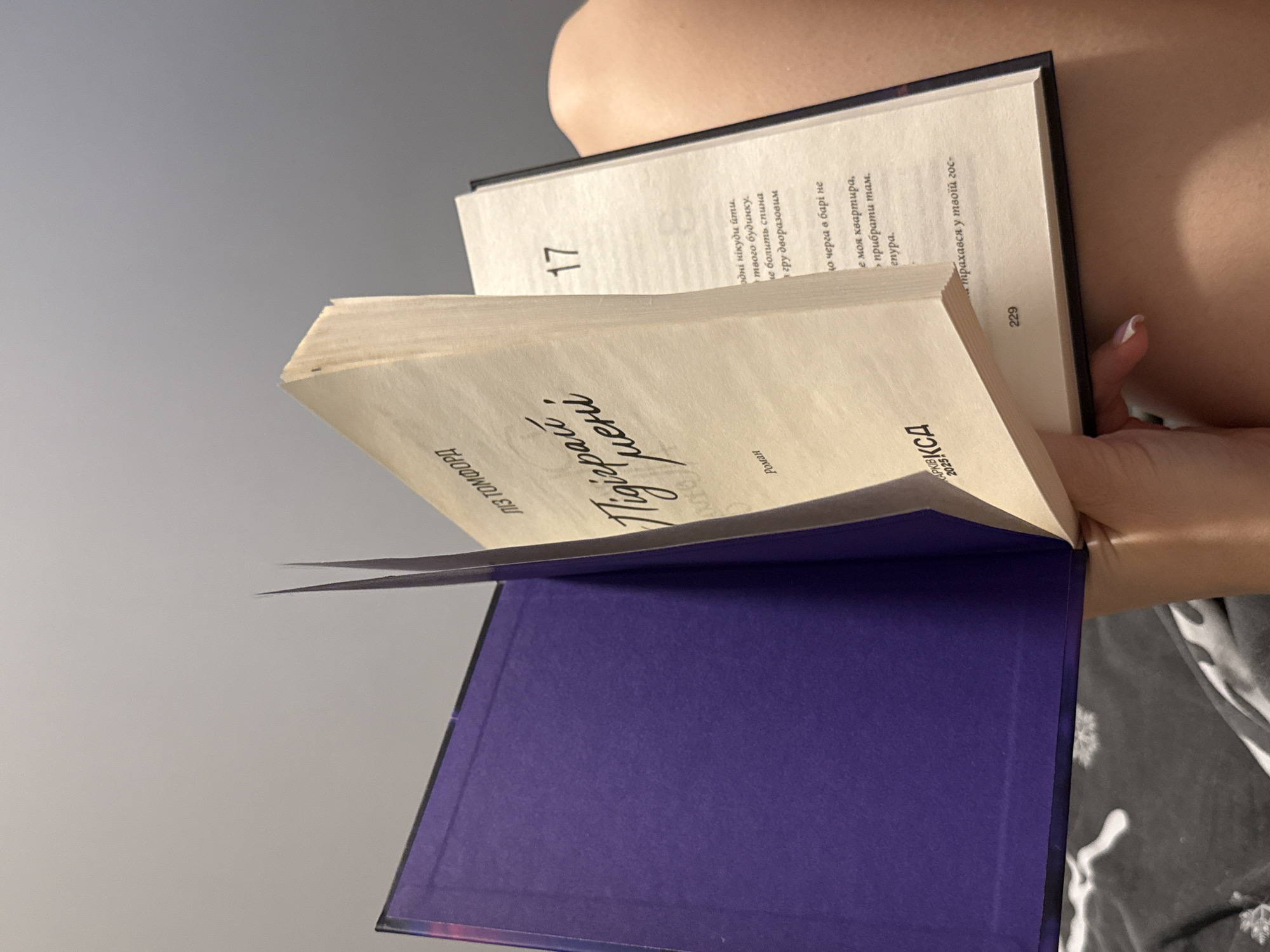 A person is holding an open book, which is angled and partially obscuring their body. The book has a purple cover on the inside, and the pages are cream-colored. The text on the pages is in Cyrillic script. On one page, the number "17" is visible, as well as some lines of text. On another page, the text "Роман ЛІЗ ТОМФОРД" is visible. The bottom of the book has text that includes "APKIB 2025". The scene appears to be indoors, with a gray background suggesting a wall or a soft light source. The person's hand, with painted fingernails, is visible holding the book. The overall impression is a candid moment of reading.