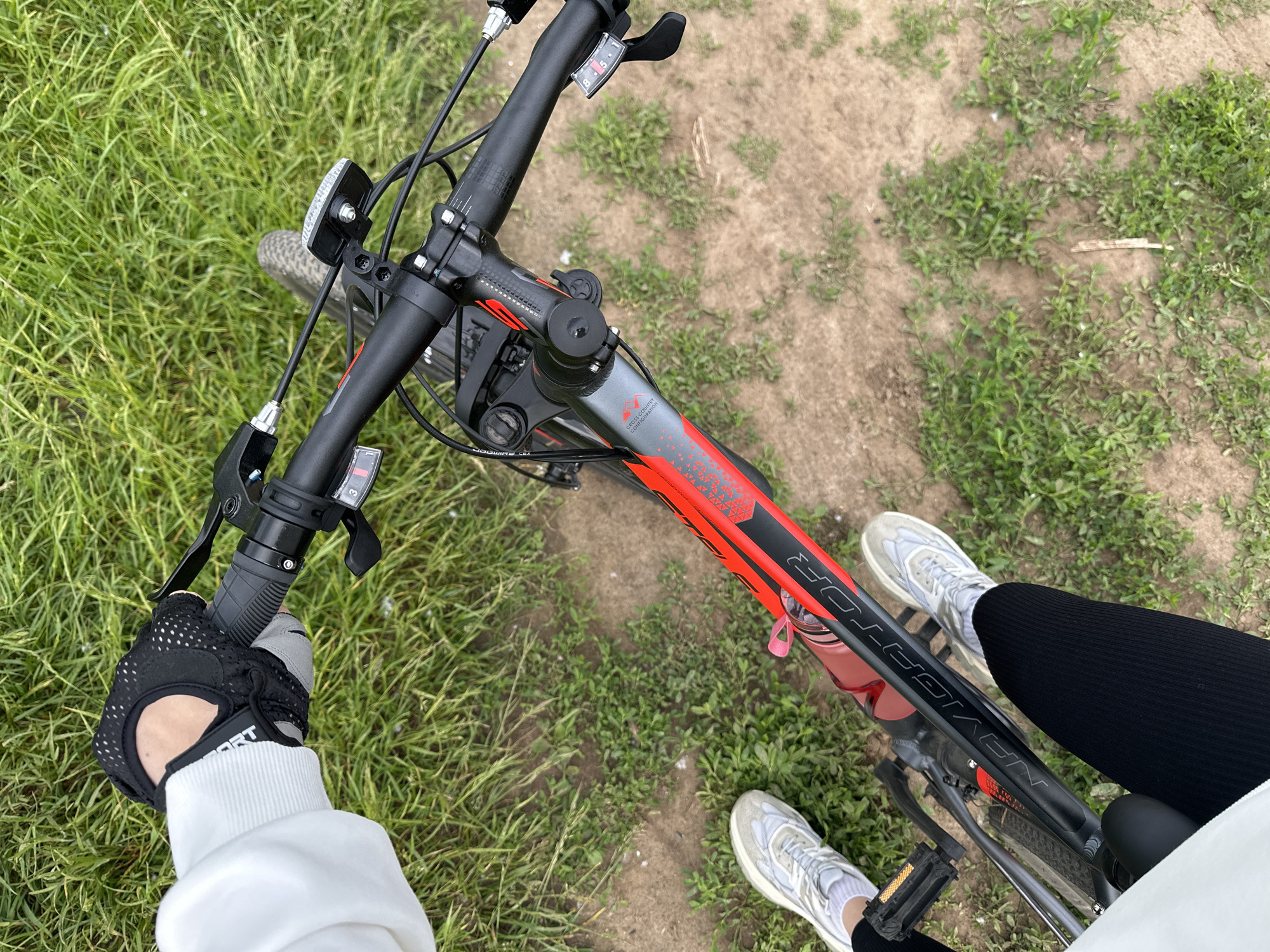 This first-person, high-angle shot captures the perspective of someone on a bicycle on an outdoor path in Chaikovsky, Russia. The rider's gloved hands, torso, and legs are partially visible. The person wears a white long-sleeved top, black fingerless cycling gloves with "SPORT" text, black leggings, and light-colored sneakers with white socks. One hand grips the left handlebar, suggesting the person is either riding or paused on the bike.
The main subject is a mountain bike featuring a black and red frame. The frame prominently displays "NAVIGATOR" in white lettering and "CROSS COUNTRY CONFIGURATION" in smaller text. A red water bottle is secured in a cage on the bike's downtube. The black handlebars are fitted with a front reflector/light, and a gear shifter showing numbers "1, 5, 6" is visible on the right side. The left grip has partially visible "JAGWIRE LTX" text. The bike appears to have knobby tires, visible at the front wheel.
The setting is an unpaved path composed of dirt and scattered green grass, transitioning to denser grass on the left and foreground. The daytime lighting indicates clear weather. The scene suggests an active outdoor pursuit, possibly cycling on a trail or park path.
