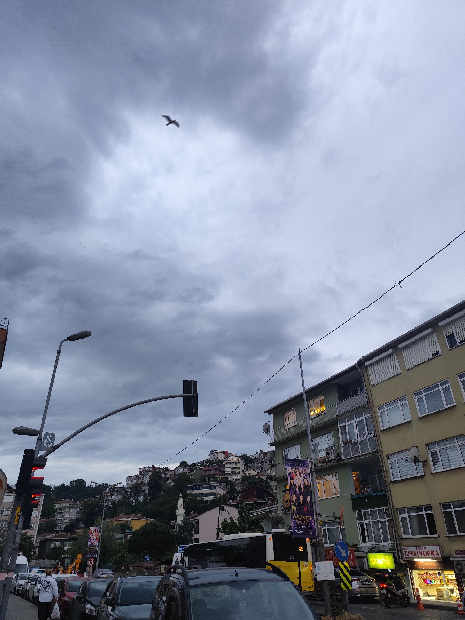 An eye-level shot displays a street scene in Sarıyer, Turkey, under a cloudy sky. A bird flies in the upper-center sky. A cluster of dwellings ascends a hillside. A light-yellow building with windows and balconies stands on the right side. A banner hangs off the side of the building. The storefront of a shop, “YUFKA YUFKA,” is lit up on the lower right. Traffic signals on the left are showing red. Several parked cars line the left side of the street. A yellow bus is behind the cars. A woman wearing a white shirt is walking on the left sidewalk. The scene appears to be a typical day in this urban area.