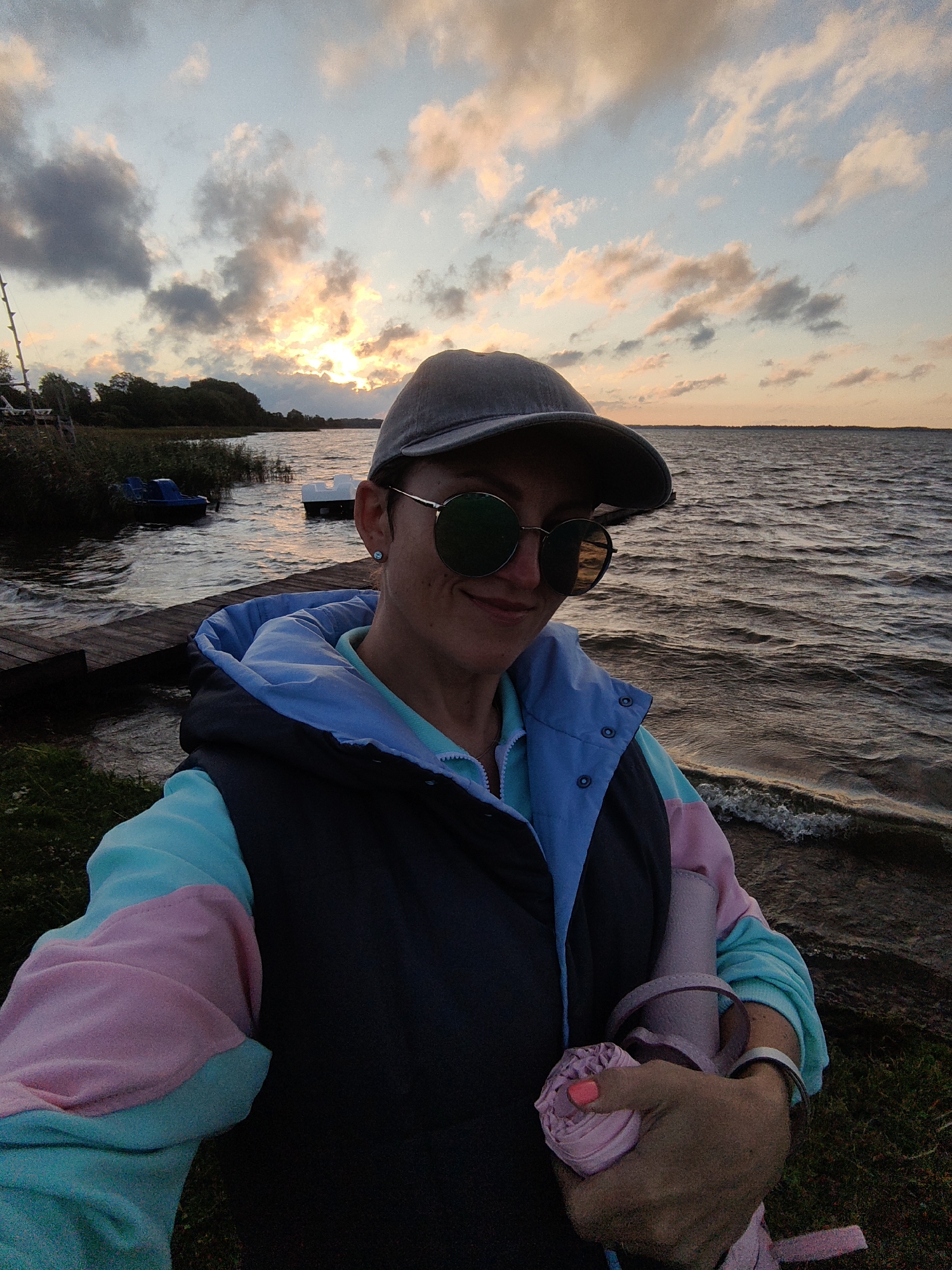 A woman wearing a grey baseball cap, round sunglasses, and a multi-colored jacket poses for a selfie by a lake at sunset in Myadzyel, Belarus. The sun is setting behind dramatic clouds, casting a warm glow across the sky and reflecting on the rippling water. A wooden dock extends into the lake, with a small boat visible in the background. The woman holds a rolled-up pink umbrella in her hand. The scene is set on the grassy bank of the lake.