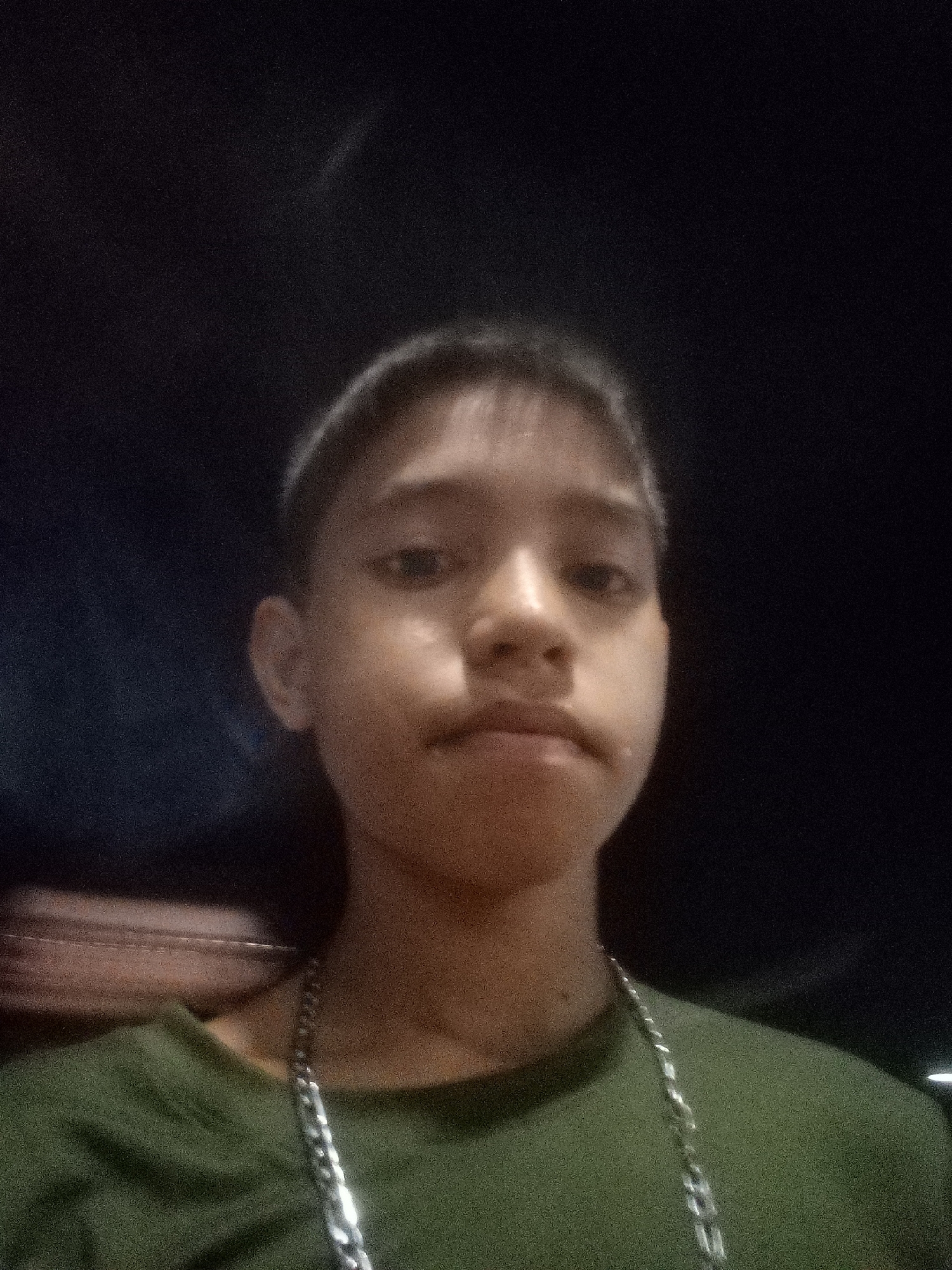 A selfie of a young Asian male with short dark hair wearing a green t-shirt and a silver chain necklace against a dark background. The image is grainy and lacking detail, potentially taken in low light. The location in Juan Griego, Venezuela cannot be determined from this image.