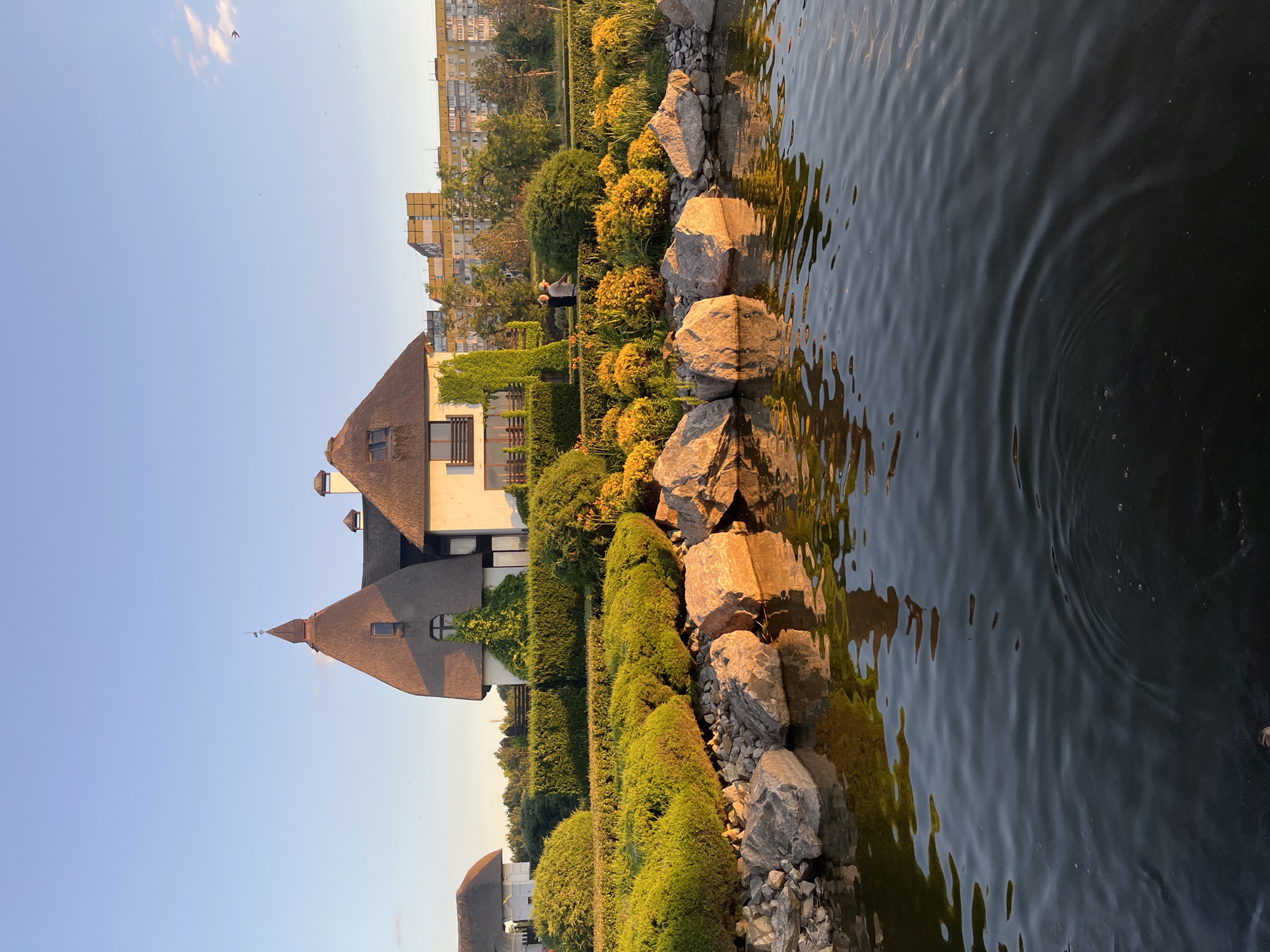 The image depicts a scenic view of a house with a thatched roof, situated in the city of Dnipro, Ukraine. The house is surrounded by a lush garden with manicured hedges and blooming yellow flowers. Large rocks are placed along the edge of a calm body of water in the foreground, reflecting the golden hues of the late afternoon sun. In the background, tall apartment buildings rise above the trees, hinting at the urban setting. Two individuals are visible in the garden, engaged in a conversation. The sky is clear blue with a few wispy clouds.