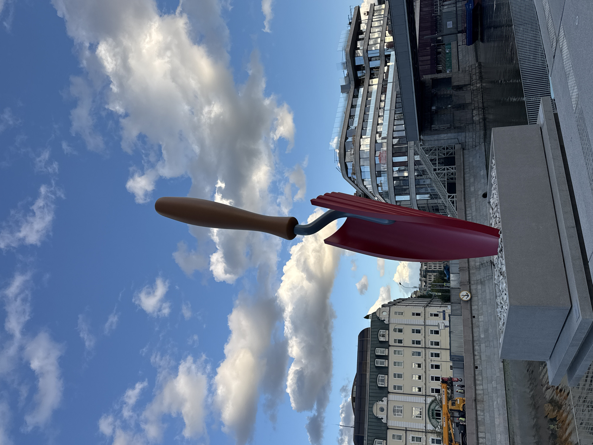 This image depicts a large, abstract sculpture of a trowel, predominantly red with a brown handle, situated on a granite pedestal filled with white stones. The sculpture is positioned outdoors, alongside a body of water, likely a river or canal. In the background, a mix of modern and older architecture is visible. To the left, a light-colored building with multiple windows and a green roof stands, with a yellow crane and several figures near it, suggesting ongoing construction or maintenance work. To the right, a contemporary building with extensive glass facades and balconies is prominent, featuring staircases leading down towards the water. A pedestrian walkway with metal railings is in the foreground. The sky above is blue with scattered white clouds. The scene appears to be a public space within a city, with elements of both historical and modern urban development. The specific location in Moscow, Russia, can be identified as being near the Moscow River embankment, potentially in the area of the Moscow International Business Center (Moscow City) or a nearby district that features this combination of architecture and the river.