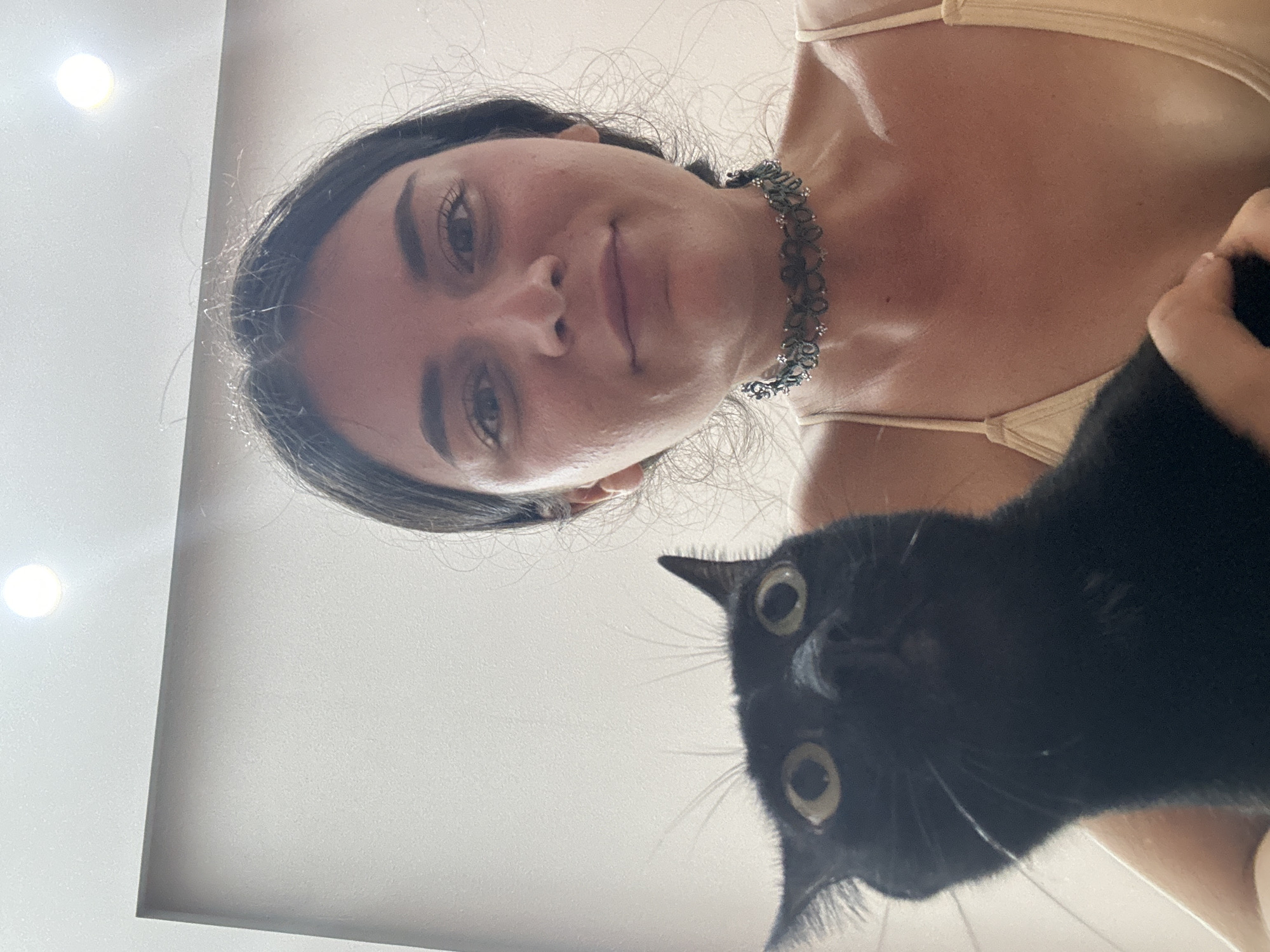 A woman with dark hair and fair skin, wearing a tan top and a floral choker, poses for a selfie with a black cat. The cat is positioned below her face, looking directly at the camera with wide eyes. The background appears to be a white, evenly lit surface. The location is not discernible from the image.