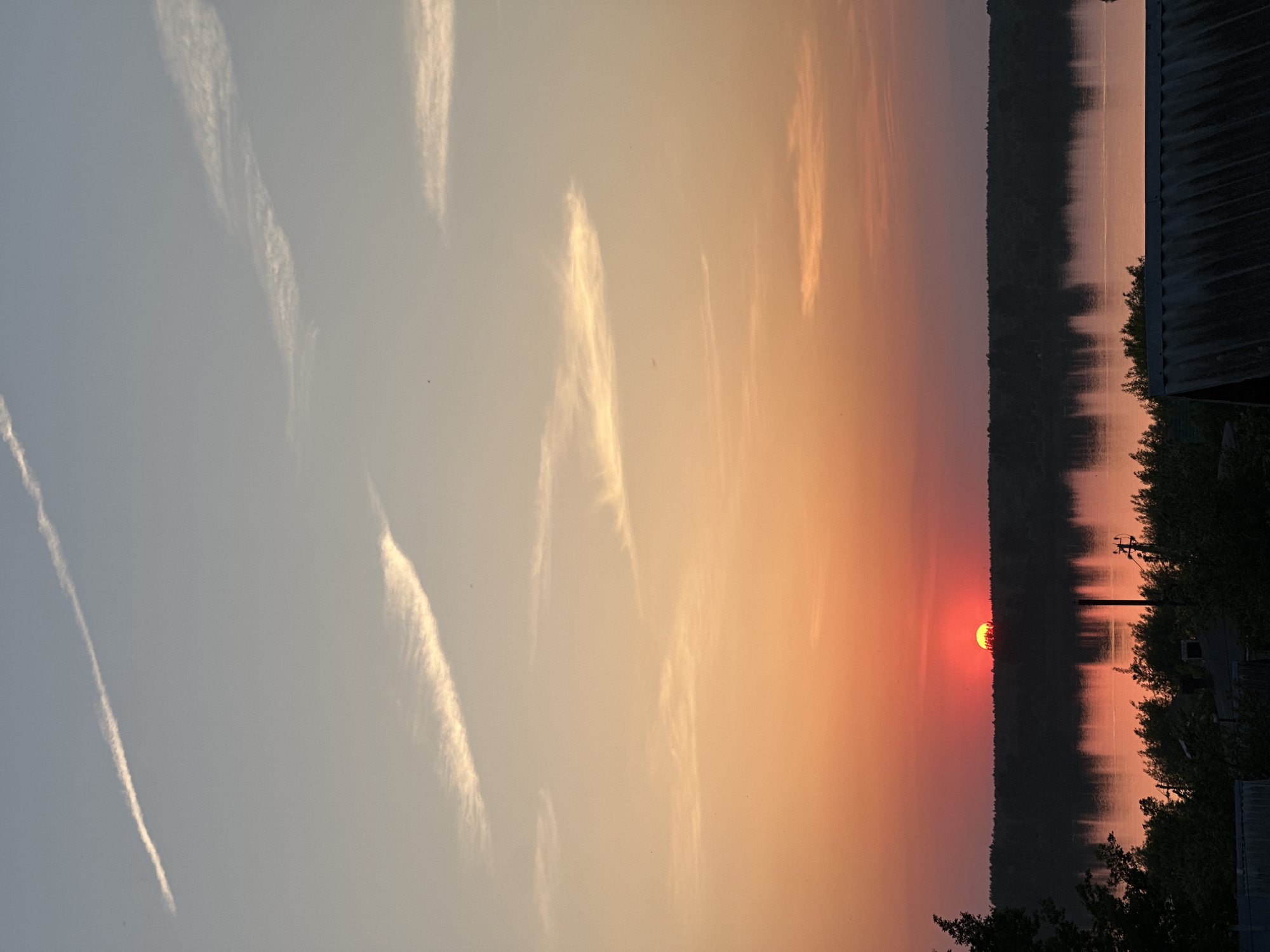 The image depicts a sunset over a body of water, likely a lake or river, in Chaikovsky, Russia. The sun is a deep red color, partially obscured by the horizon, which is lined with trees. The sky transitions from a light blue in the upper left to a deep orange near the horizon. Several contrails are visible in the sky. The water reflects the colors of the sunset. On the right side of the frame, foliage and what appears to be a building with a dark roof are visible. An electrical pole is partially visible among the trees.
