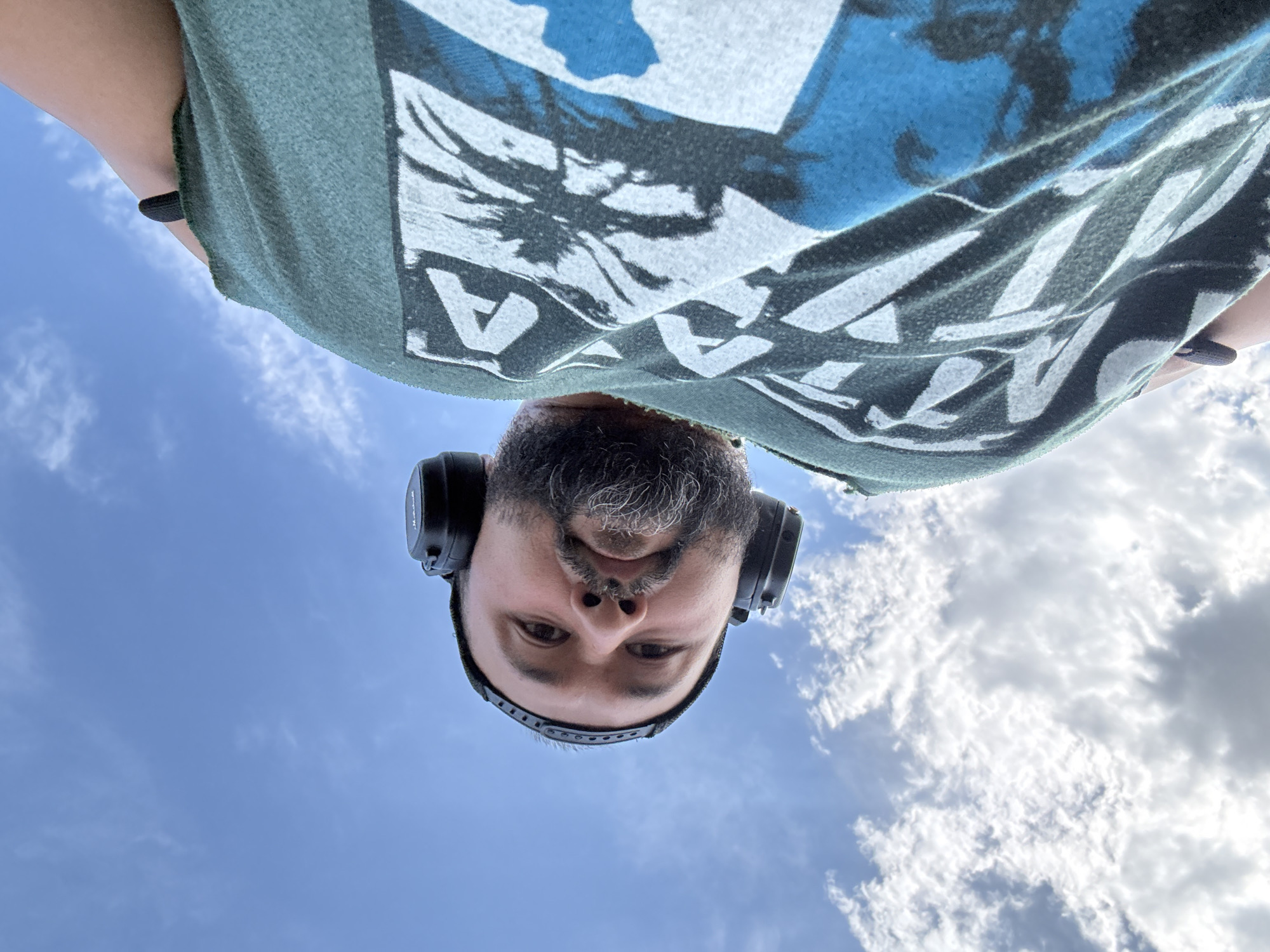 This is a low-angle, wide-shot, outdoor, close-up photograph of a man wearing headphones. The man is looking up at the camera, and his face is partially obscured by his beard. He is wearing a green t-shirt with a graphic design on it. The background is a bright blue sky with scattered white clouds. The image does not provide any specific information about the location within the city of Ақши, Kazakhstan.