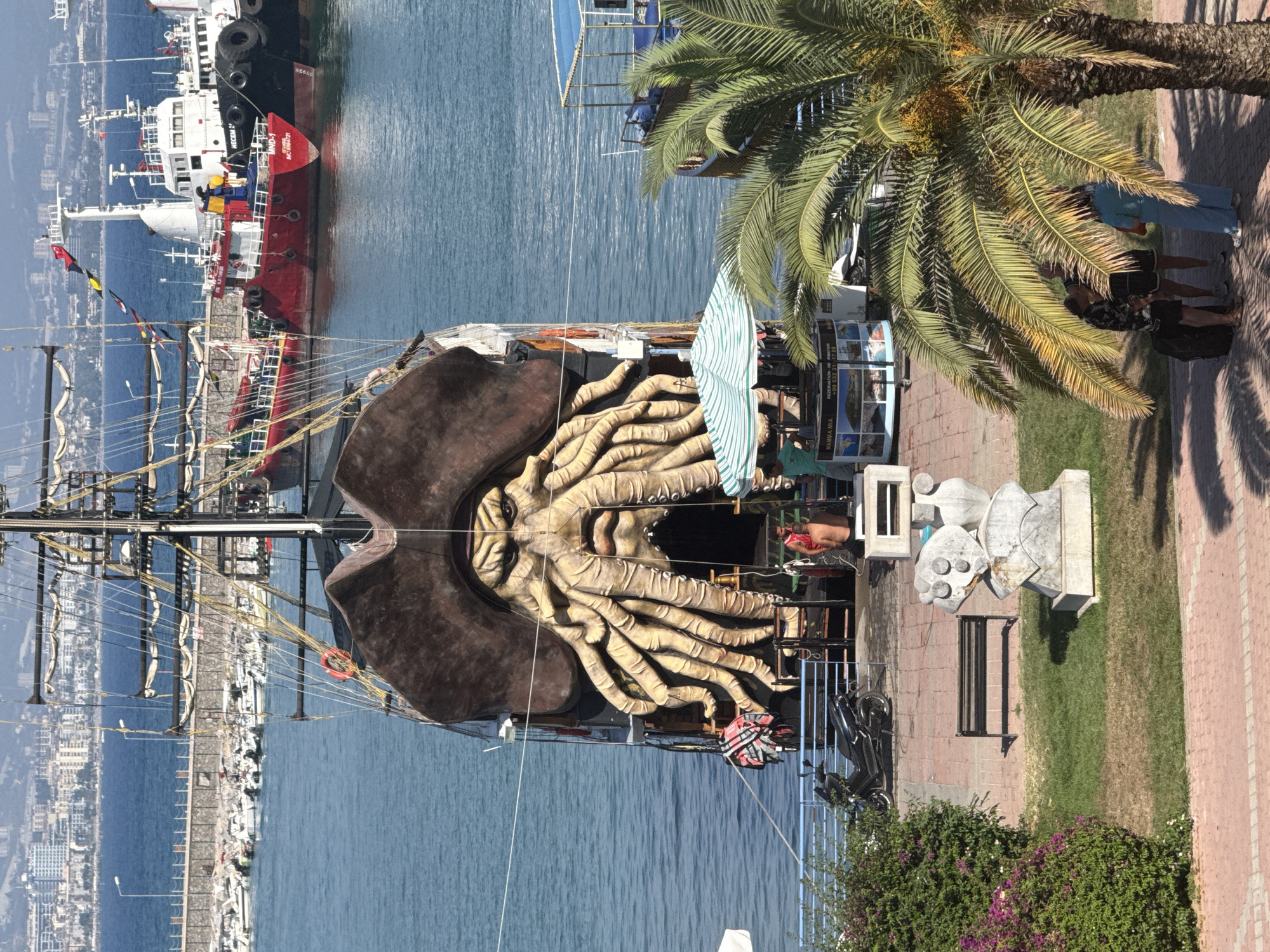 The image captures a scenic view of the Alanya harbor in Turkey, with a prominent pirate-themed ship docked in the foreground. The ship features a large, imposing figurehead resembling a monstrous face with tentacles, designed to resemble Davy Jones from Pirates of the Caribbean. In the background, other vessels are visible, including a large red tugboat and a white ship, along with a city skyline.
In the immediate foreground, a paved walkway leads to a grassy area with a park bench and a modern sculpture. A palm tree with its fronds casting shadows adds to the tropical ambiance. Several people are scattered throughout the scene:
* Two individuals are standing near the palm tree, appearing to converse.
* Another person, wearing a blue outfit, is walking away from the palm tree, carrying a bag.
* Near the pirate ship, two men are standing. One, partially visible, is shirtless and appears to be looking at the ship. The other is wearing a green shirt and is interacting with a kiosk.
* A few other figures are obscured by the ship's structure or distant boats.
Other objects in the scene include a black scooter parked on the walkway and a striped umbrella providing shade over a kiosk. The water is calm and reflects the sunlight, suggesting a clear day. The overall impression is that of a bustling tourist location, showcasing elements of both maritime activity and leisure.