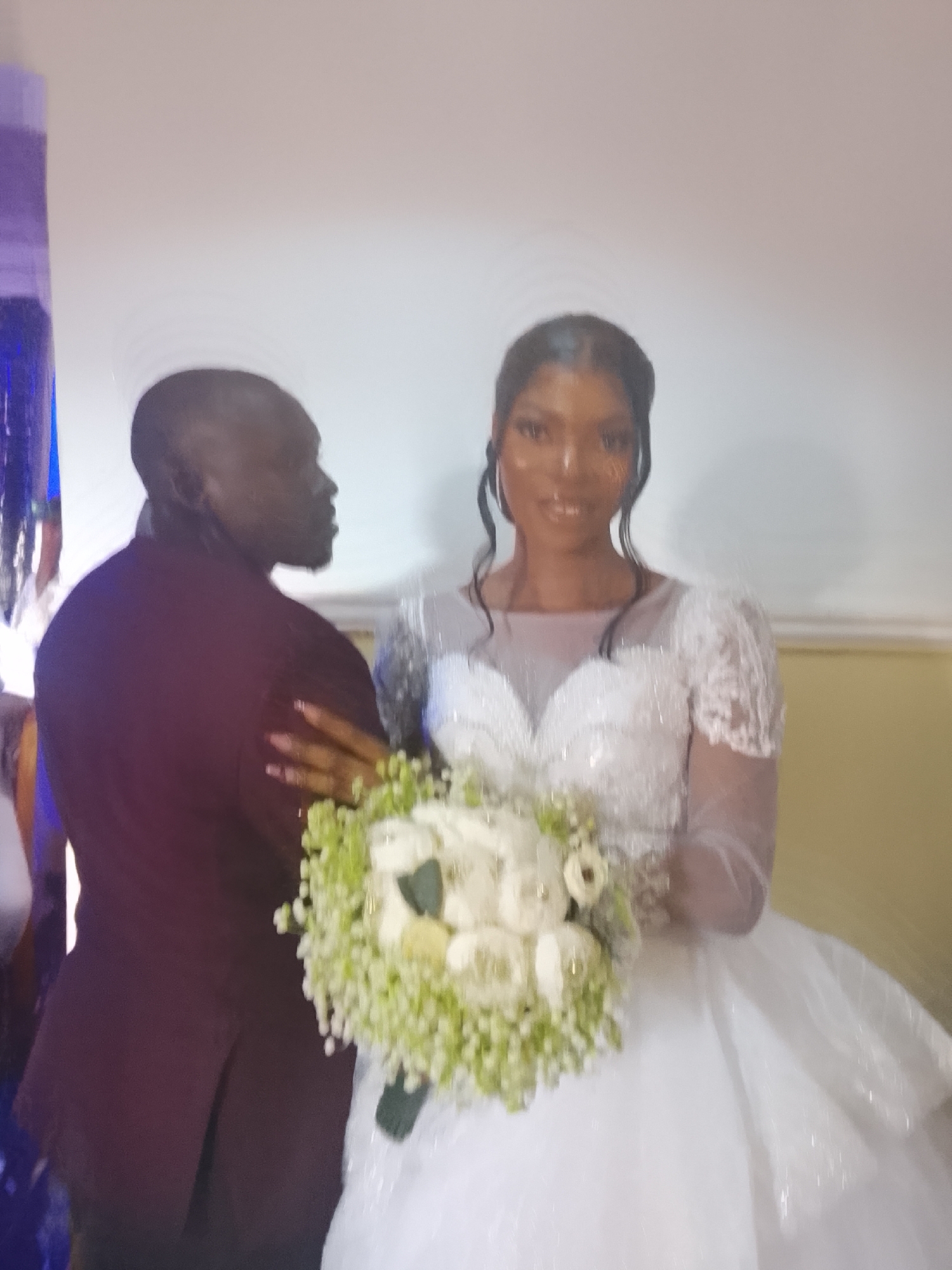 The image shows a wedding in an indoor setting. A bride in a white gown is holding a bouquet of white flowers with green accents. A man in a maroon jacket stands beside her, looking to the side. The location of the wedding is likely in the city of Dogon Dinia, Nigeria.