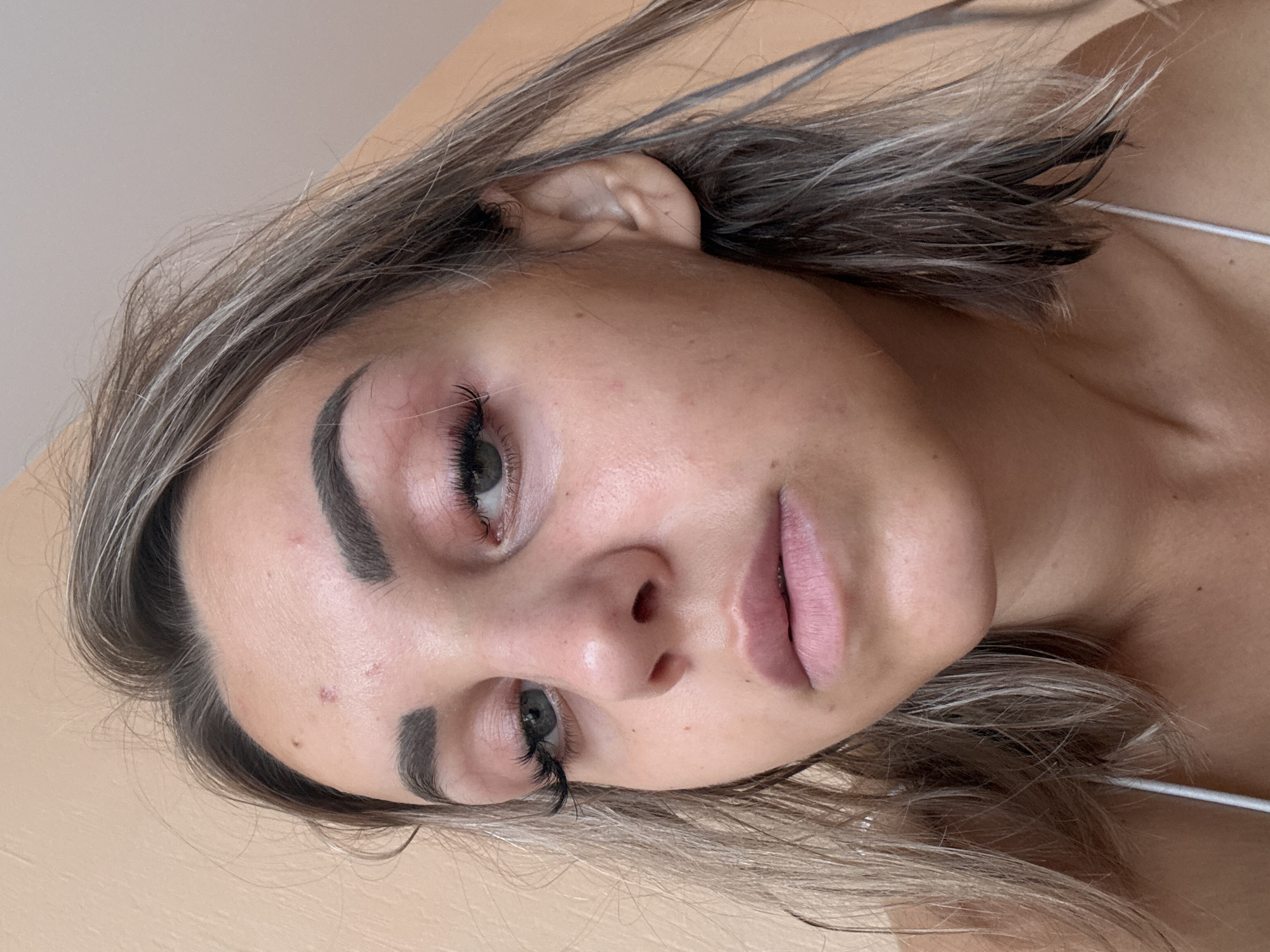 A close-up, eye-level, natural light portrait shows the face of a young woman with blond hair that is layered and appears damp. Her eyes are looking slightly to the left, and she has on false eyelashes. Her skin has some redness around her right eye, and there are a few small blemishes on her forehead. She is wearing a white tank top. The background is a plain, light-colored wall. The image does not contain any specific indicators of location in Kholmsk, Russia, or any other geographical place.