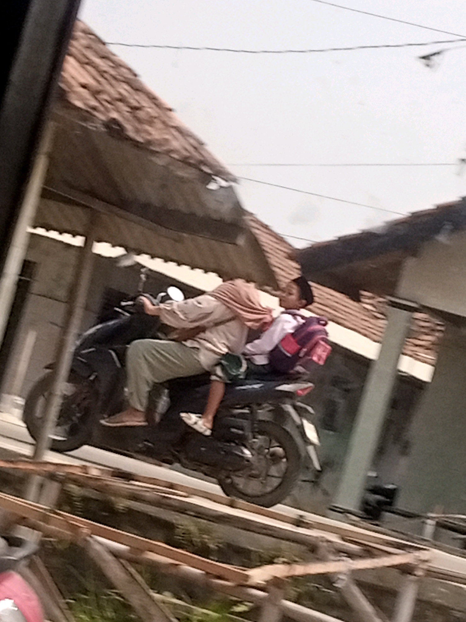 In Boyoteluk, Indonesia, two individuals are riding on a dark-colored motorcycle. The person in the rear, a child wearing a black cap and a white shirt with a pink backpack, is perched on the back of the motorcycle. The person in the front, presumed to be an adult, is wearing a light-colored shirt and light-colored pants, with a headscarf covering their hair. The motorcycle is traveling on an unpaved road, with a traditional Indonesian tiled roof house and a building with an overhang visible in the background. The sky is overcast.