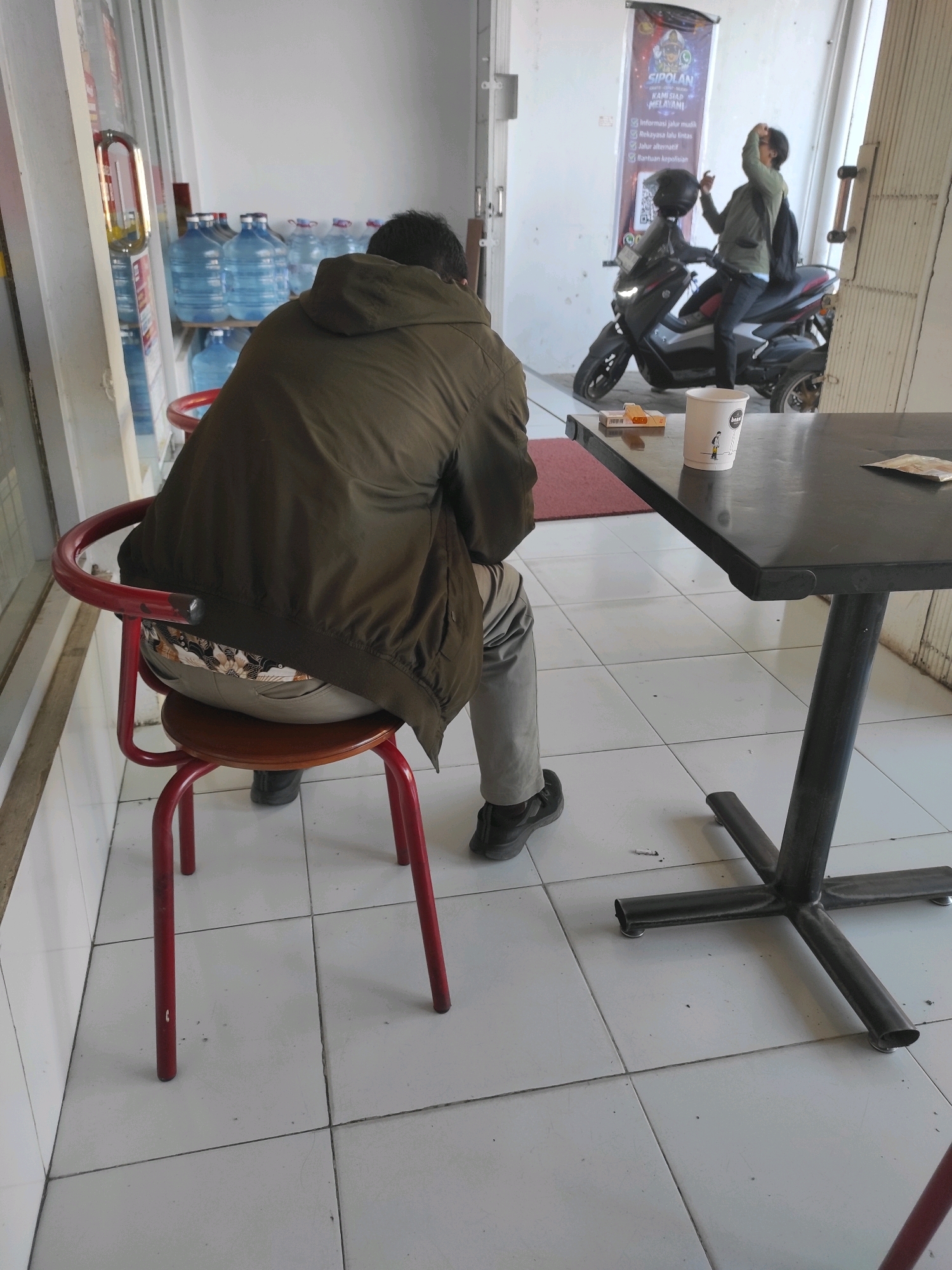 In Purwodadi, Indonesia, a man wearing a dark green hooded jacket and light gray pants sits on a red metal-framed stool. His back is to the camera, and his head is turned to the right, suggesting he is looking towards a motorcycle with two people on it parked outside. The motorcycle is black with red accents. One person on the motorcycle, wearing a green jacket, is looking down and holding a phone up as if taking a photo or video. The other person on the motorcycle is obscured. In the foreground, a black table stands with a white paper cup, a pack of cigarettes, and a lighter on it. The floor is tiled in a light color. In the background, blue water dispenser bottles are stacked on shelves. The scene appears to be in an outdoor or semi-outdoor commercial space, possibly a cafe or rest stop.