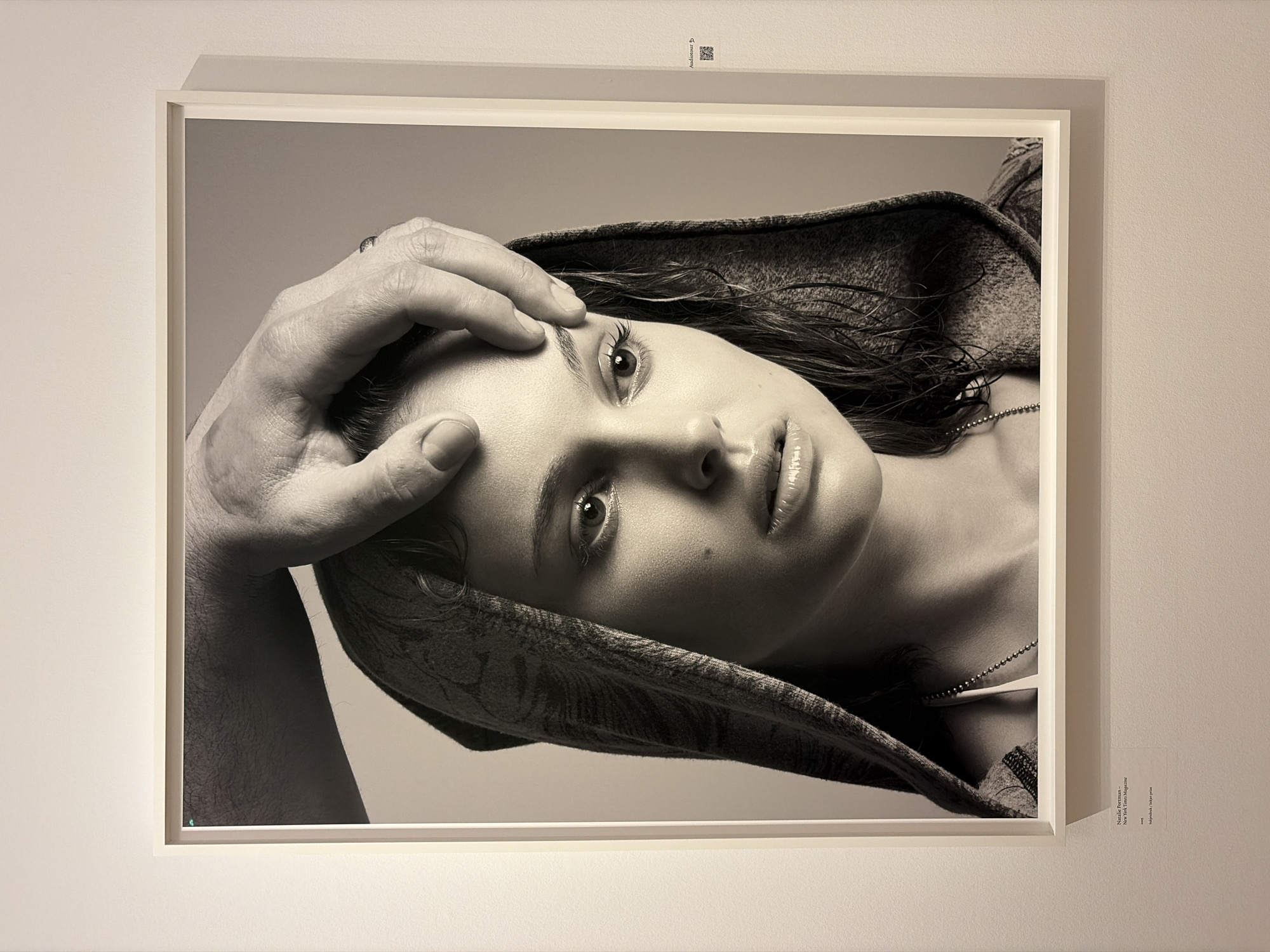 The image shows a black-and-white framed photograph displayed on a light-colored wall. The photograph features a close-up portrait of a woman with dark, wet-looking hair, wearing what appears to be a patterned head covering or shawl. A man's hand rests on the woman's forehead, his arm visible on the left side of the frame. The portrait is in a white frame with a white mat. Above the photograph on the wall is a small sign with an audioguide symbol and QR code. To the right of the photograph is a label with text, identifying the artwork as "Nadja Portrait, New York, NY Magazine" by Jurgen Teller, and noting that it is inkjet print. The photograph is at the Fotomuseum in The Hague, Netherlands.