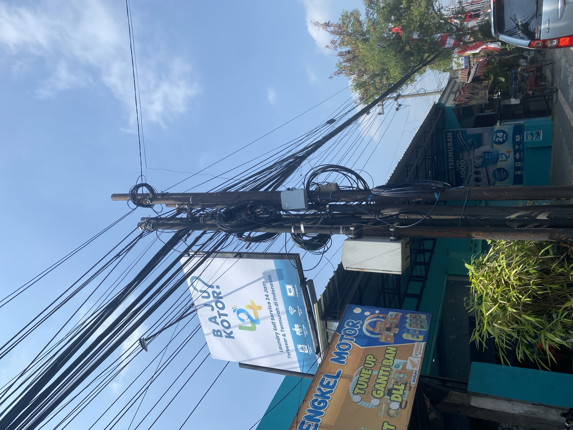A street scene in Kediri City, Indonesia, is dominated by a utility pole laden with a dense network of black electrical and telecommunications wires, casting a stark contrast against a bright blue sky with sparse white clouds. A prominent white billboard for "Baju Kotor!" (Dirty Clothes!) laundry service hangs from the pole, advertising "Laundry Self Service 24 Jam Terbesar & Termurah di Indonesia" (Largest & Cheapest 24 Hour Self Service Laundry in Indonesia) and displaying icons for washing machines, dryers, high-speed Wi-Fi, and a 4K TV. Below this, another sign for "ENGKEL MOTOR" (Motorcycle Workshop) is visible, indicating automotive services are offered. Adjacent to the utility pole, a turquoise-colored building with a covered walkway houses the "LAUNDRY TERMURAH" (Cheapest Laundry) business. This laundry has its own signage detailing services and pricing, including "CUMA 1000/KG" (Only 1000/KG) and "Datang - Cuci - Kering - Puas" (Come - Wash - Dry - Satisfied). Further down the street, a silver car is parked on the right, and Indonesian flags are displayed on buildings, suggesting a national holiday or celebration. Lush green plants, including bamboo, grow near the building, adding a natural element to the urban environment. The overall impression is a busy commercial street with essential services catering to the local community.