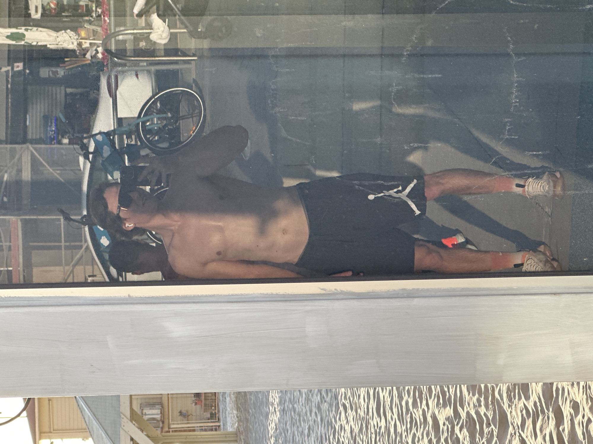 The image, oriented correctly, depicts a detailed reflection of a man lying down in a large glass surface, likely a storefront window, during daytime. The man is shirtless, wearing dark shorts adorned with a white Jumpman logo on the left thigh, and white sneakers. He has sunglasses on and is holding a smartphone to his face, possibly capturing a selfie or viewing content. His torso appears toned, and his skin is tanned, with a noticeable sunburn (redness) on his lower legs.
Reflected in the glass alongside the man is a bicycle, partially visible, suggesting an outdoor urban setting such as a street or sidewalk. The glass surface shows visible dust and streaks.
Through the glass, the background reveals an indoor or covered area with industrial-looking structures, possibly metal shelving or frameworks. At the bottom of the image, below a white window frame, there's a textured surface resembling sand dunes or rippled sand, illuminated by sunlight. The scene conveys a casual, sunny atmosphere, aligning with the Tel-Aviv, Israel context.