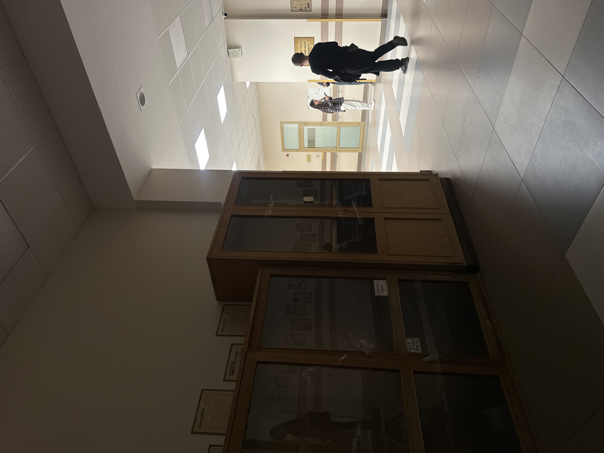 The image depicts a corridor in Kazan, Russia. A man in a dark outfit is walking down the hallway with another person in the background, possibly with their phone in hand, while another person stands near the door. The corridor has tile flooring with a geometric design and is lined with display cabinets containing documents and artifacts. The lighting appears to be fluorescent from the ceiling, which has a grid of tiles.