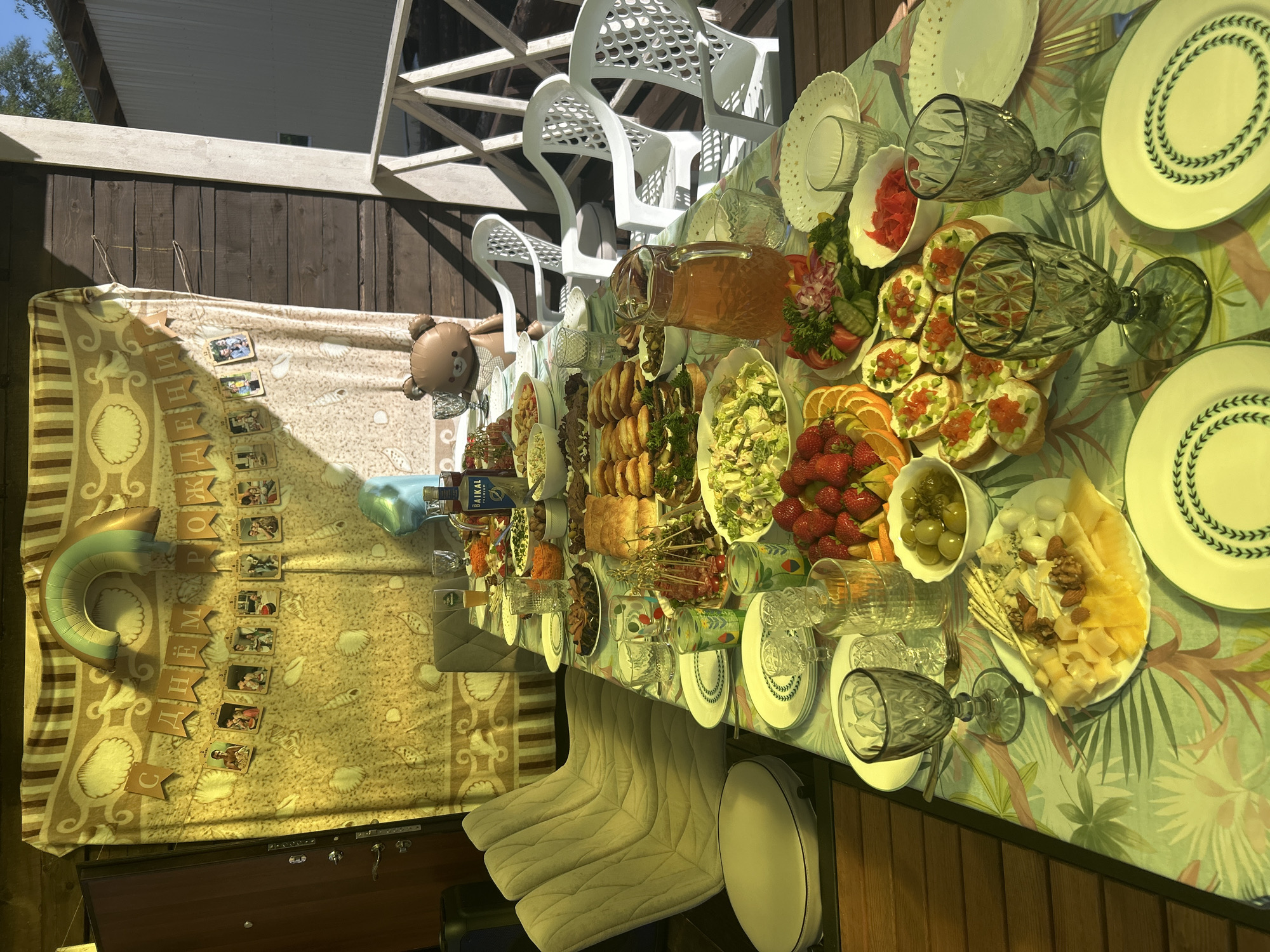 The image shows an outdoor dining table set up on a wooden porch, likely in Lyamino, Russia, during a celebration. A patterned tablecloth covers the table, which is laden with a variety of food and drinks. Visible items include plates, glasses, a pitcher of orange liquid, several bowls of food (some containing olives, strawberries, and what appears to be salad and appetizers), a platter of cheeses and nuts, and several small sandwiches with toppings. In the background, a draped fabric with a shell pattern serves as a backdrop, decorated with a banner that reads "С ДНЕМ РОЖДЕНИЯ" (Happy Birthday) and a string of photographs. A large balloon in the shape of a rainbow and a small inflatable teddy bear are also present. Behind the table and chairs, wooden railings and the side of a building are visible, suggesting a residential or recreational setting. The lighting indicates it is daytime, with sunlight casting shadows on the scene. There are no people clearly visible in the image.