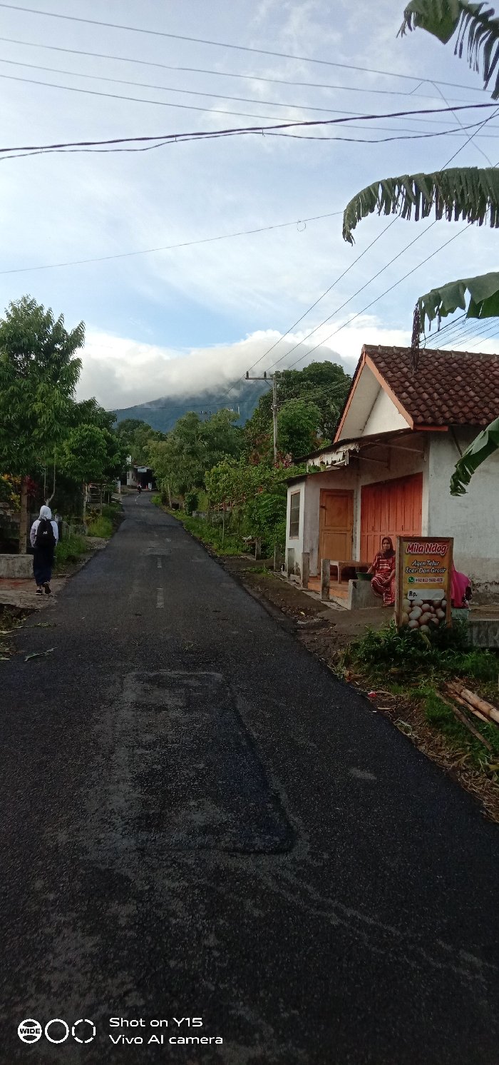 This vertical shot captures a peaceful, semi-rural street scene in Ranugedang, Indonesia, likely in the early morning or late afternoon given the soft light and cloudy sky. A winding asphalt road, with some patches and a faint white line, recedes into the distance, leading towards lush green mountains partially obscured by low-hanging clouds.
On the left side of the road, a person wearing a white hoodie and a black backpack walks away from the camera. The road is flanked by dense tropical foliage, including large trees. On the right, a simple, single-story building with a white facade, an orange door, and a brown tiled roof stands prominently. A woman in a patterned dress is seated on a wooden bench in front of this building, looking towards the road. Behind her, partially obscured, is another person.
Attached to the building is a brown sign advertising "Mila Ndog," identifying it as an "Agen Telur Ecer Dan Grosir" (Egg Agent, Retail and Wholesale), with a phone number "+62 812 7080-6019" and "Rp." visible. Overhead, power lines crisscross the bright, cloudy sky, with large, dry banana leaves hanging into the frame on the right. The overall atmosphere is calm and appears to depict everyday life in a village with a scenic mountain backdrop. A "Shot on Y15 Vivo AI camera" watermark is present in the bottom left corner.